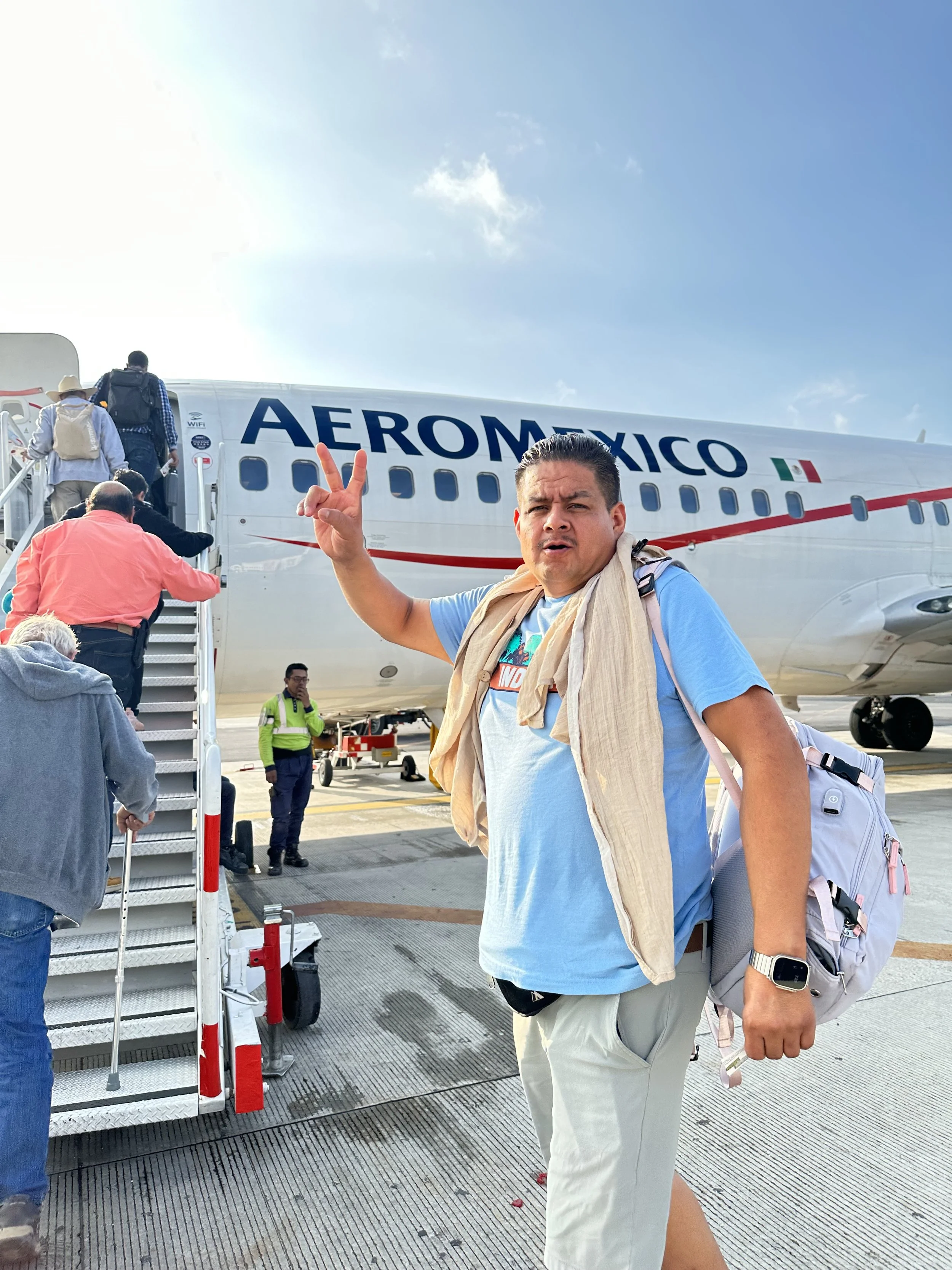 Person giving a peace sign in front of an AeroMexico airplane on the tarmac with other travelers boarding the plane.