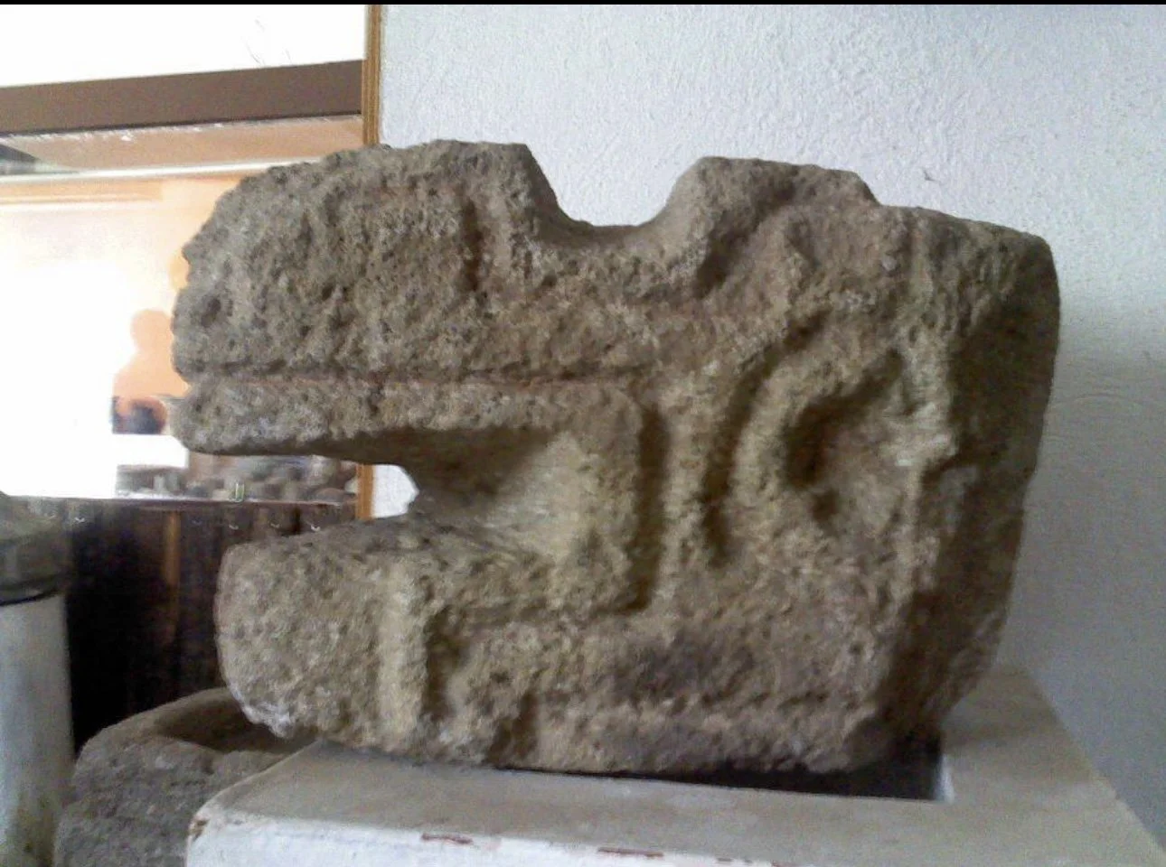 Ancient stone carving featuring a humanoid face, displayed indoors on a white pedestal.