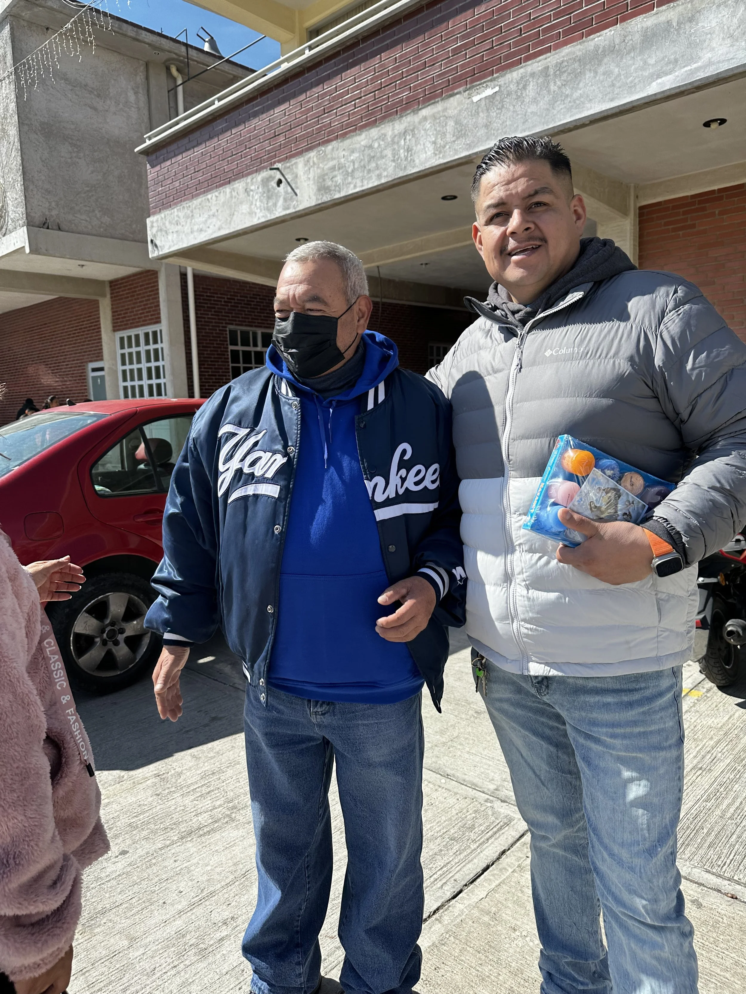 Two men standing outdoors in front of a red car and a brick building. The man on the left wears a blue Yankees jacket and a black face mask. The man on the right wears a gray jacket and holds a bag of small toys.