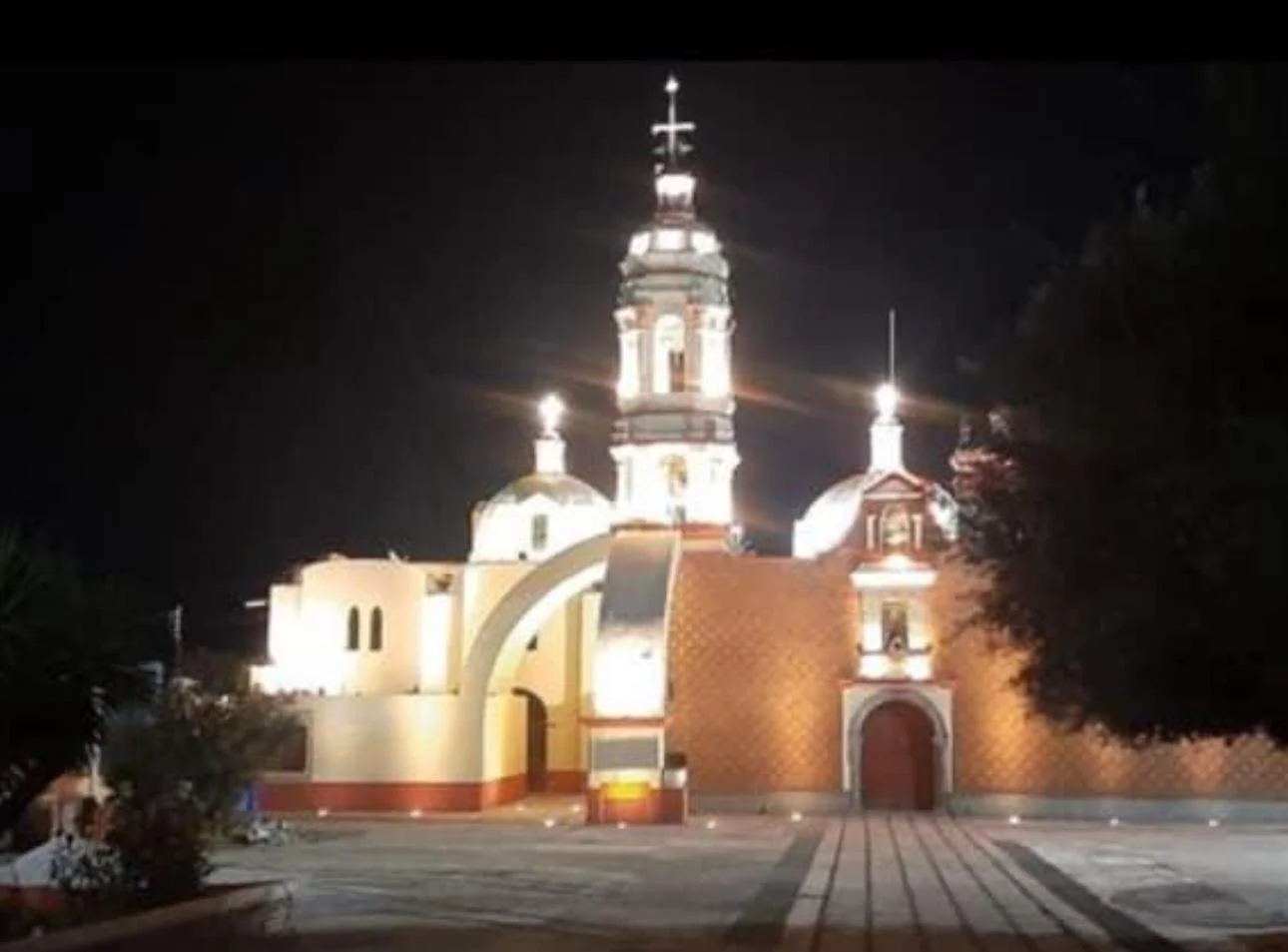 Night view of a church with bright exterior lights, featuring a tall central tower topped with a cross, and two smaller domes with crosses, a decorative brick wall, and an arched entrance.