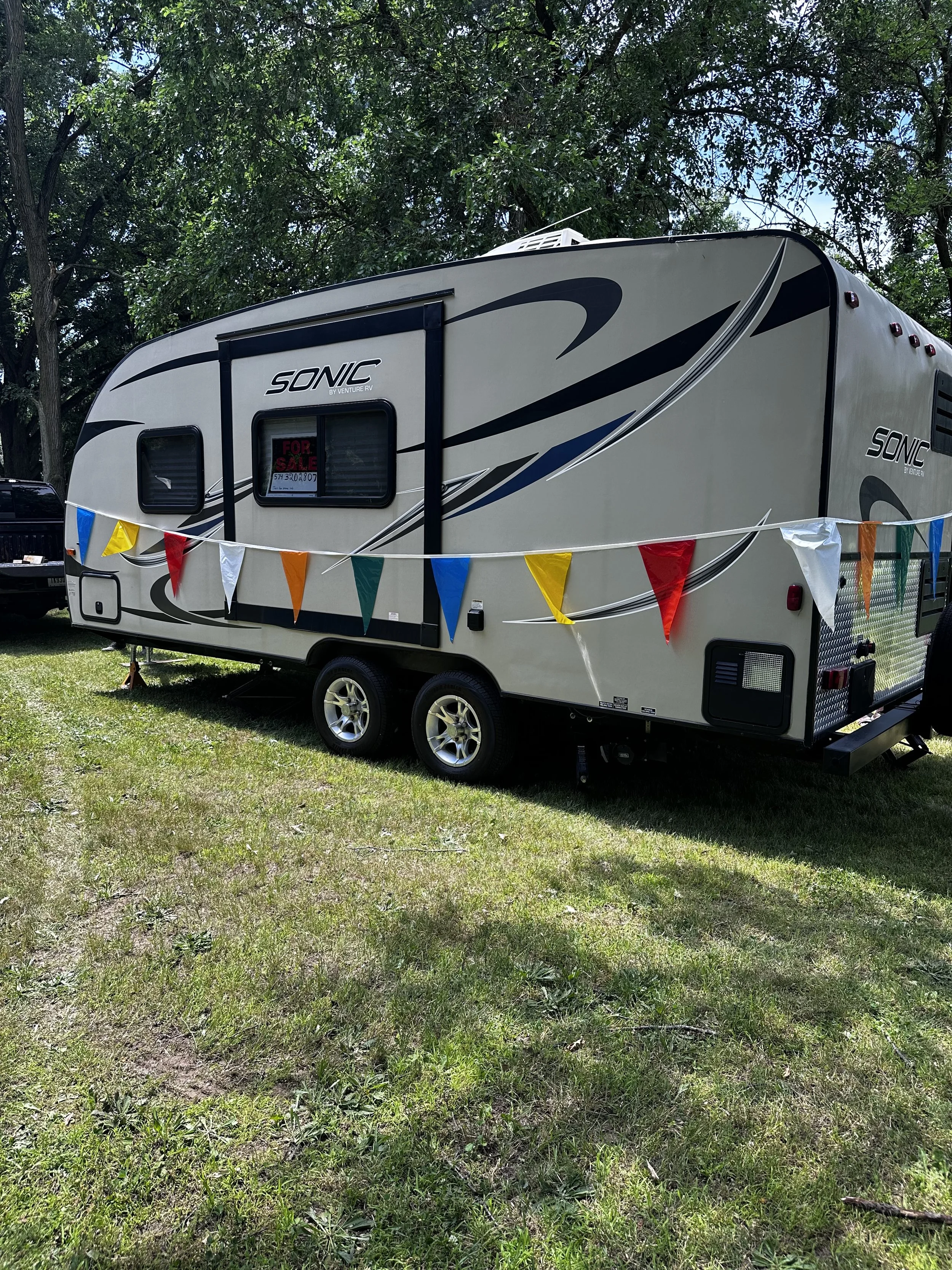 A beige Sonic RV trailer decorated with multicolored flags, parked on grass under trees with a for sale sign in the window.
