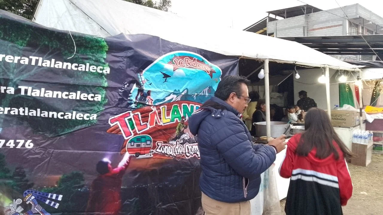 Two men standing close together indoors. The younger man on the left wears a light blue polo shirt and beige pants, has keys hanging from his pocket. The older man on the right wears glasses, a purple and brown argyle sweater over a collared shirt. They appear to be engaged in a conversation.