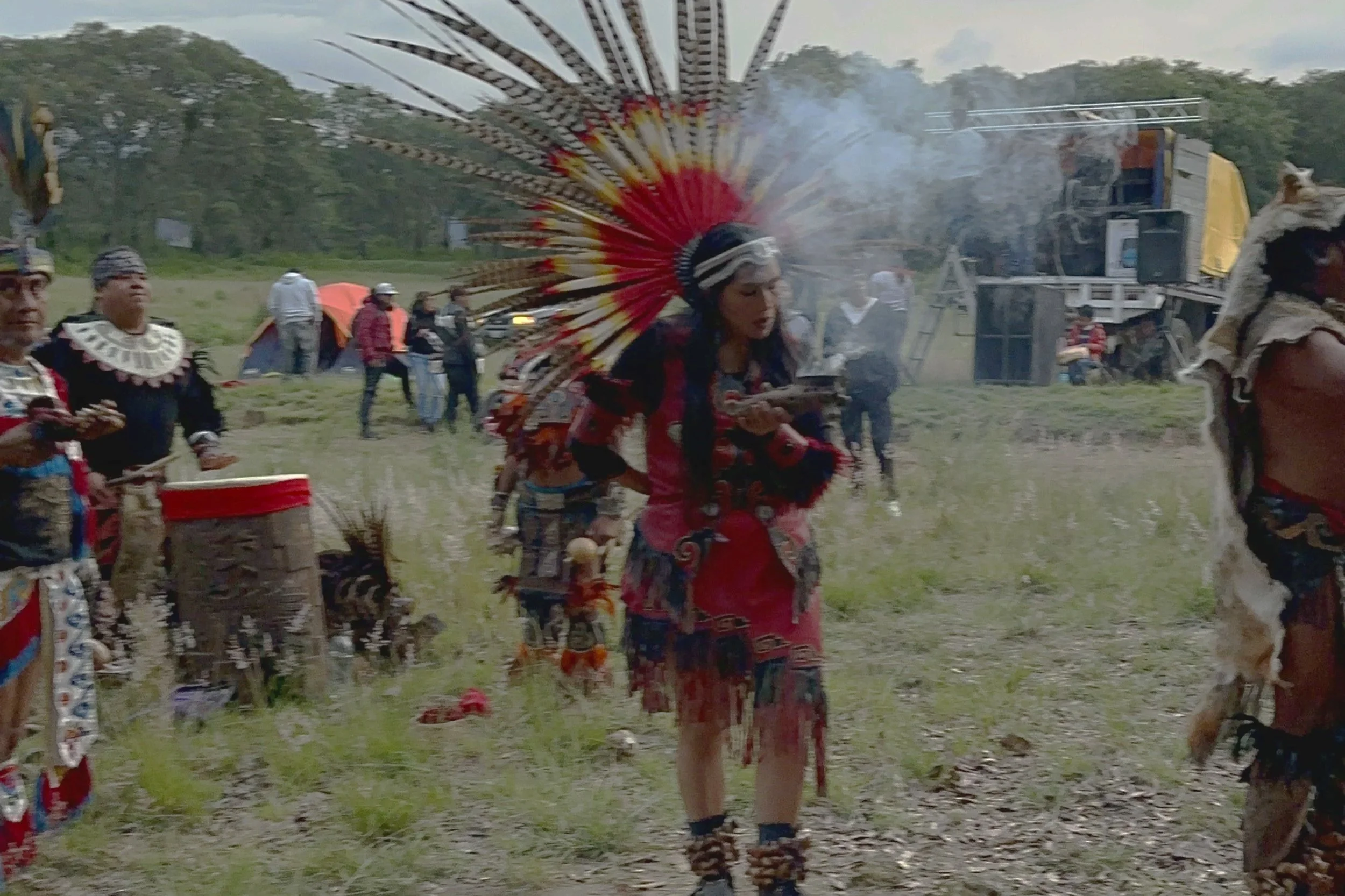 Indigenous people in traditional attire participating in a cultural event outdoors, with one woman wearing a feathered headdress, holding a plate, and others playing drums and wearing face paint.