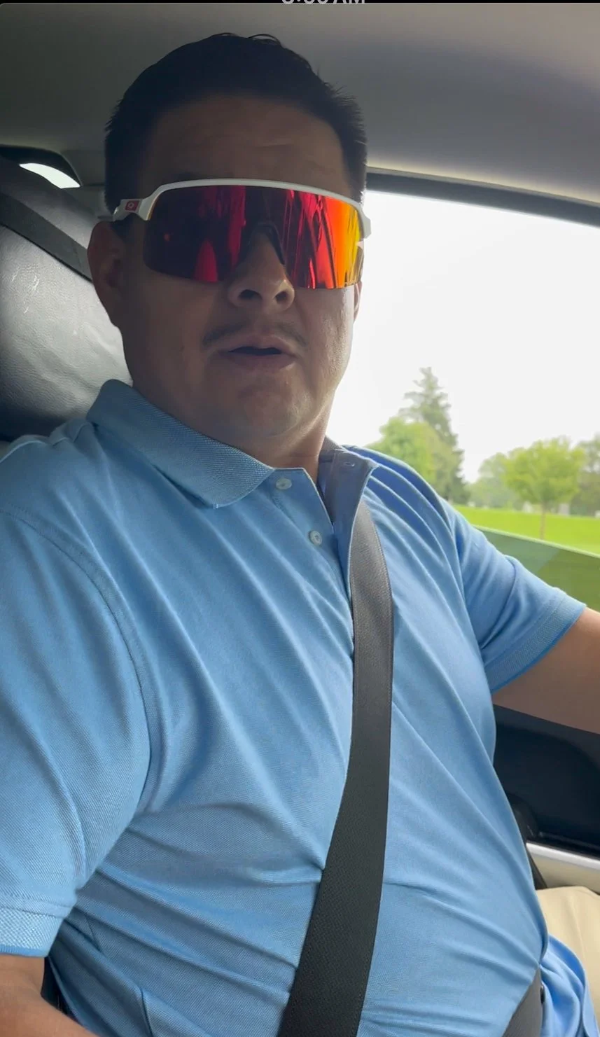 A man wearing a blue polo shirt and red-tinted sports sunglasses sitting in a car with trees and a bright sky visible through the window.