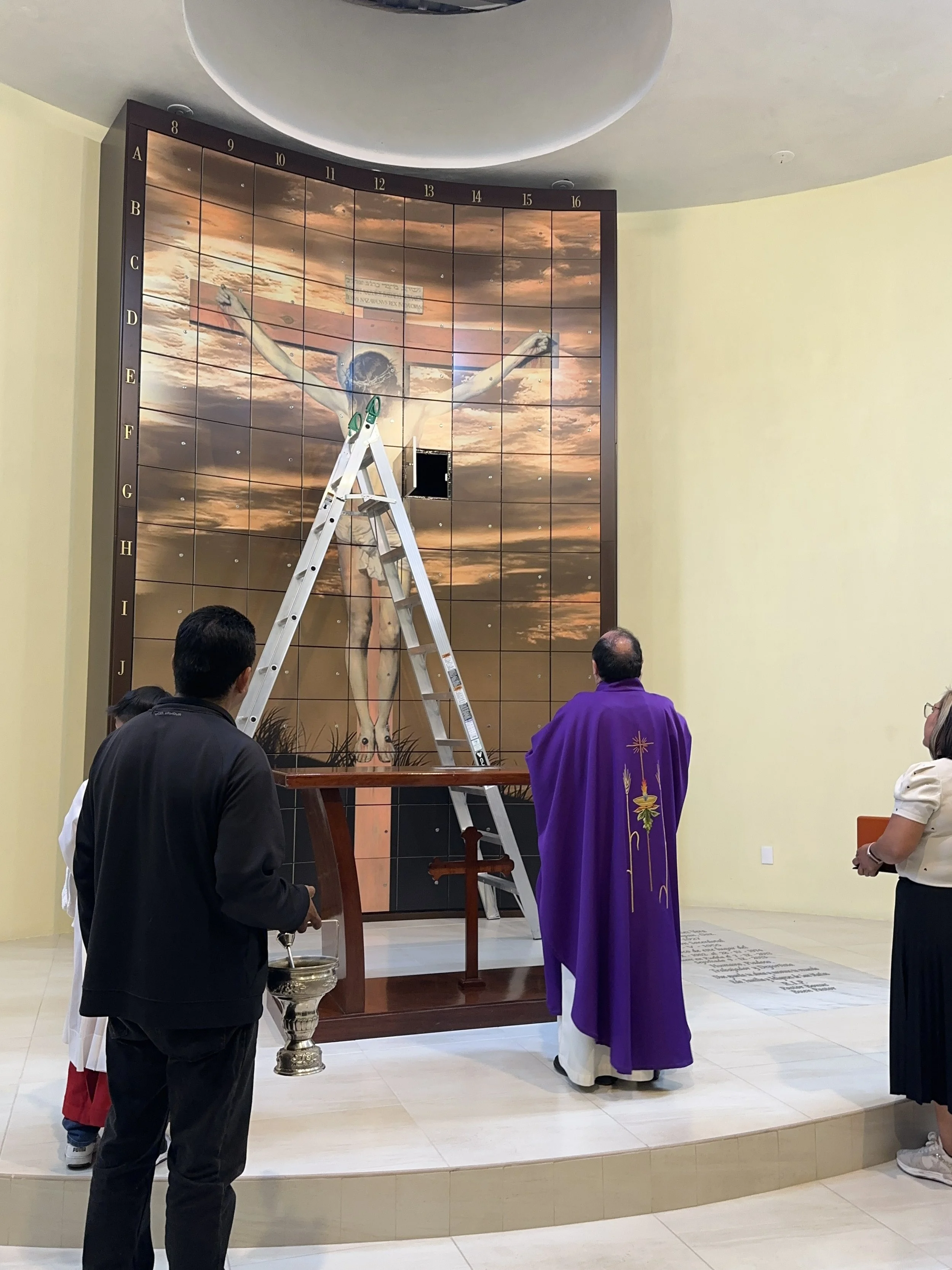 People standing in front of a large religious mural of Jesus on the cross, with a ladder placed against the mural, in a church