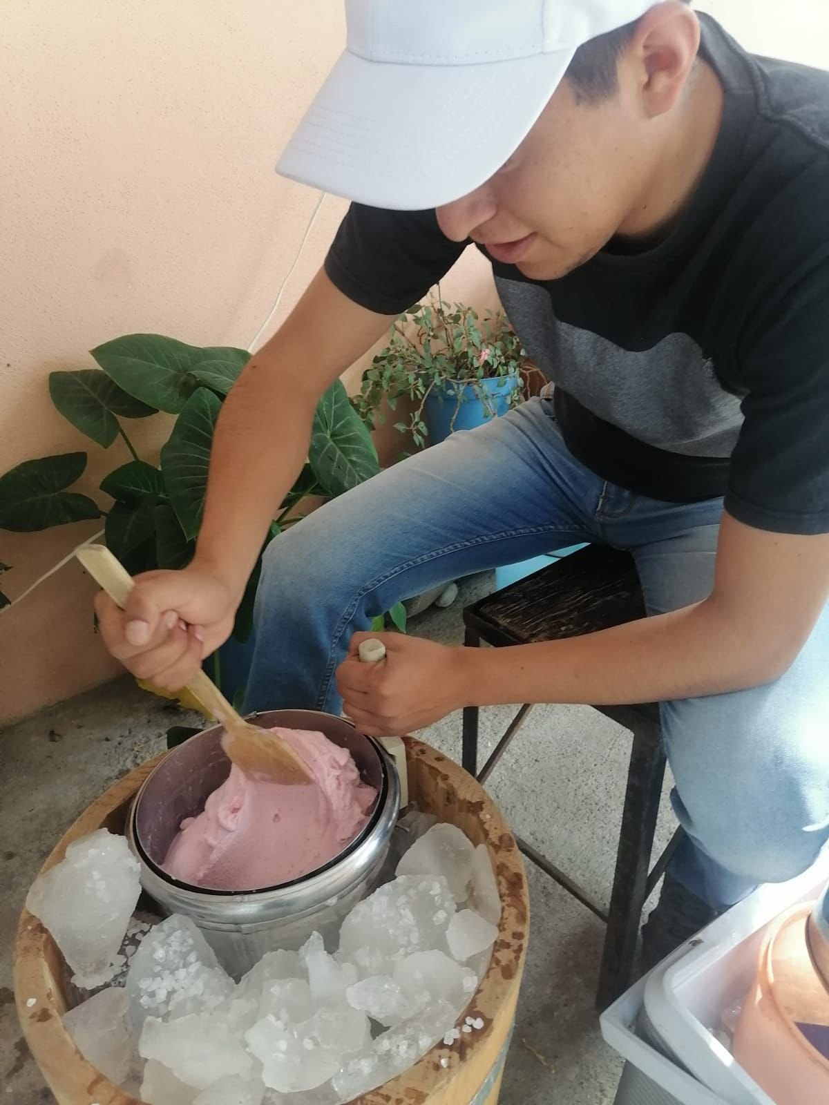 A person wearing a white cap and black T-shirt, sitting on a stool, stirring pink ice cream in a bowl, which is placed on a wooden table surrounded by ice blocks.