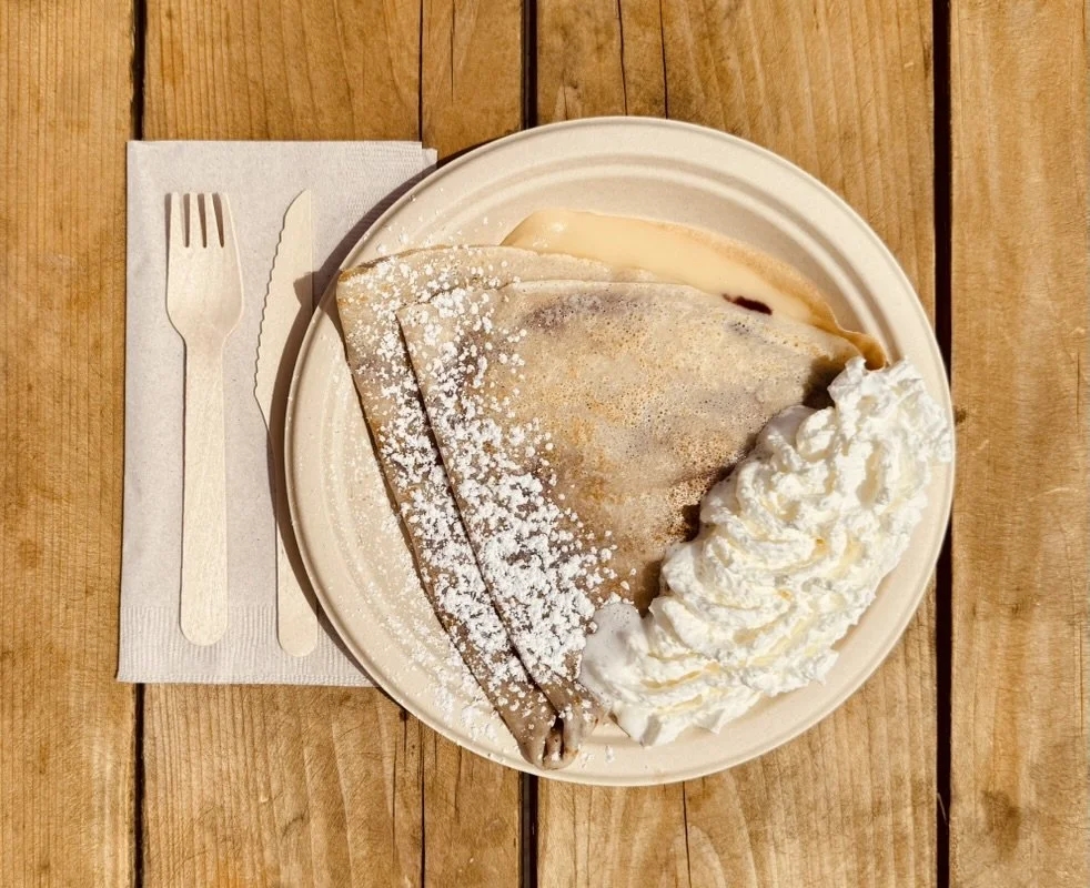A slice of crepe cake with whipped cream on a white plate, dusted with powdered sugar, served with a side of whipped cream. The plate is on a wooden table with a paper napkin, fork, and knife.