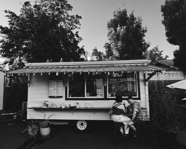 A small food truck with a sliding window serving area, parked outdoors surrounded by trees and plants, with two people sitting on a bench in front, talking and looking at a phone.