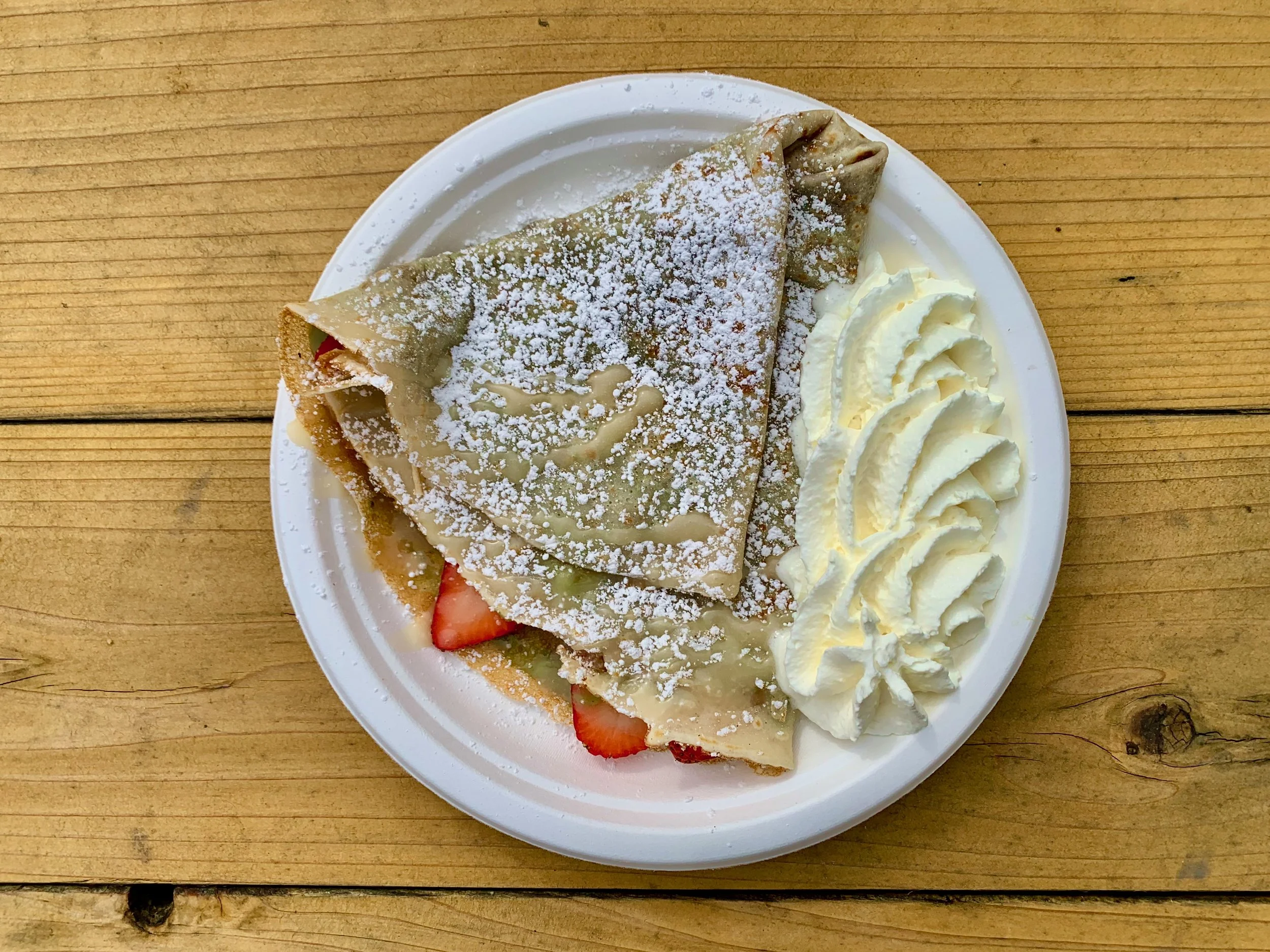 A plate with a folded crepe sprinkled with powdered sugar, served with whipped cream and slices of strawberries and other fruits on a wooden table.