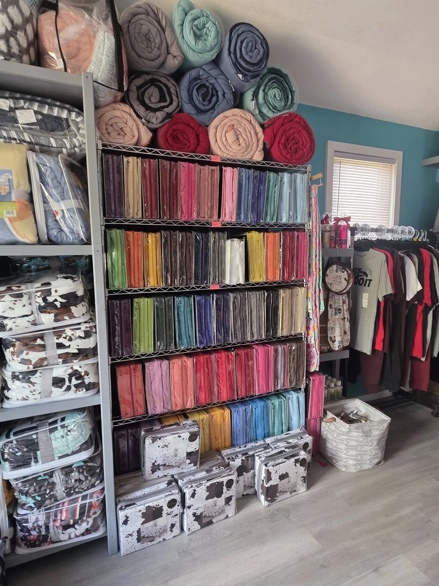 Shelving unit with rolls of fabric in various colors, including pink, green, blue, and purple, with some white and black patterned fabric and boxes at the bottom, in a craft or fabric store.