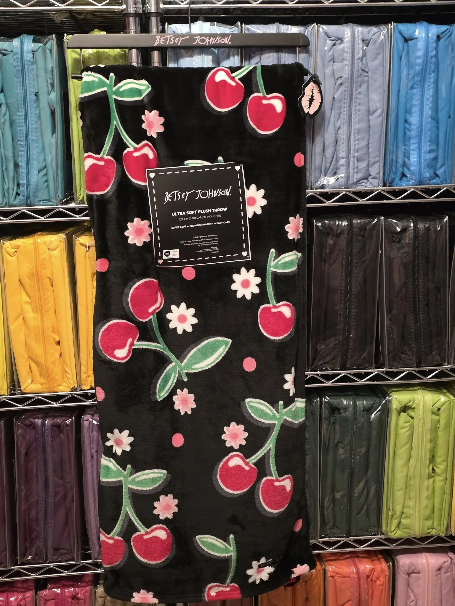 Black plush throw blanket with cherry and flower pattern hanging on a retail display, surrounded by colorful zippered luggage in a store.