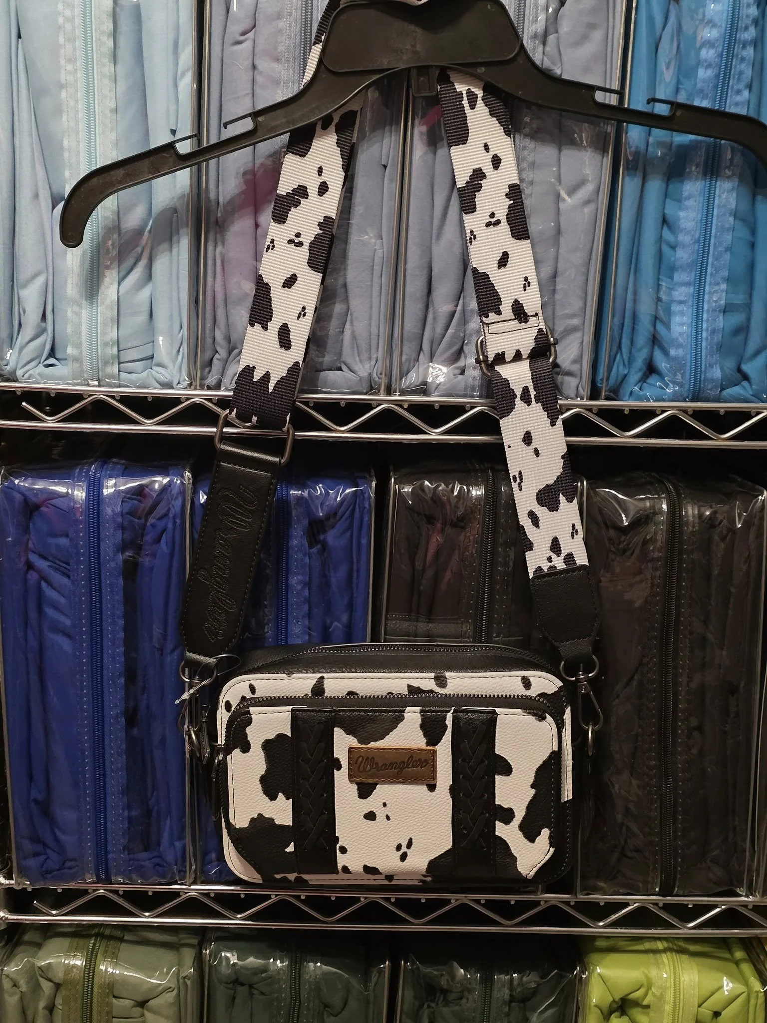 A black and white cow print crossbody bag hanging on a plastic coat hanger in front of a shelf of folded travel pouches. The bag has a leather patch with 'Wrangler' embossed on it, and a wide, cow print strap.