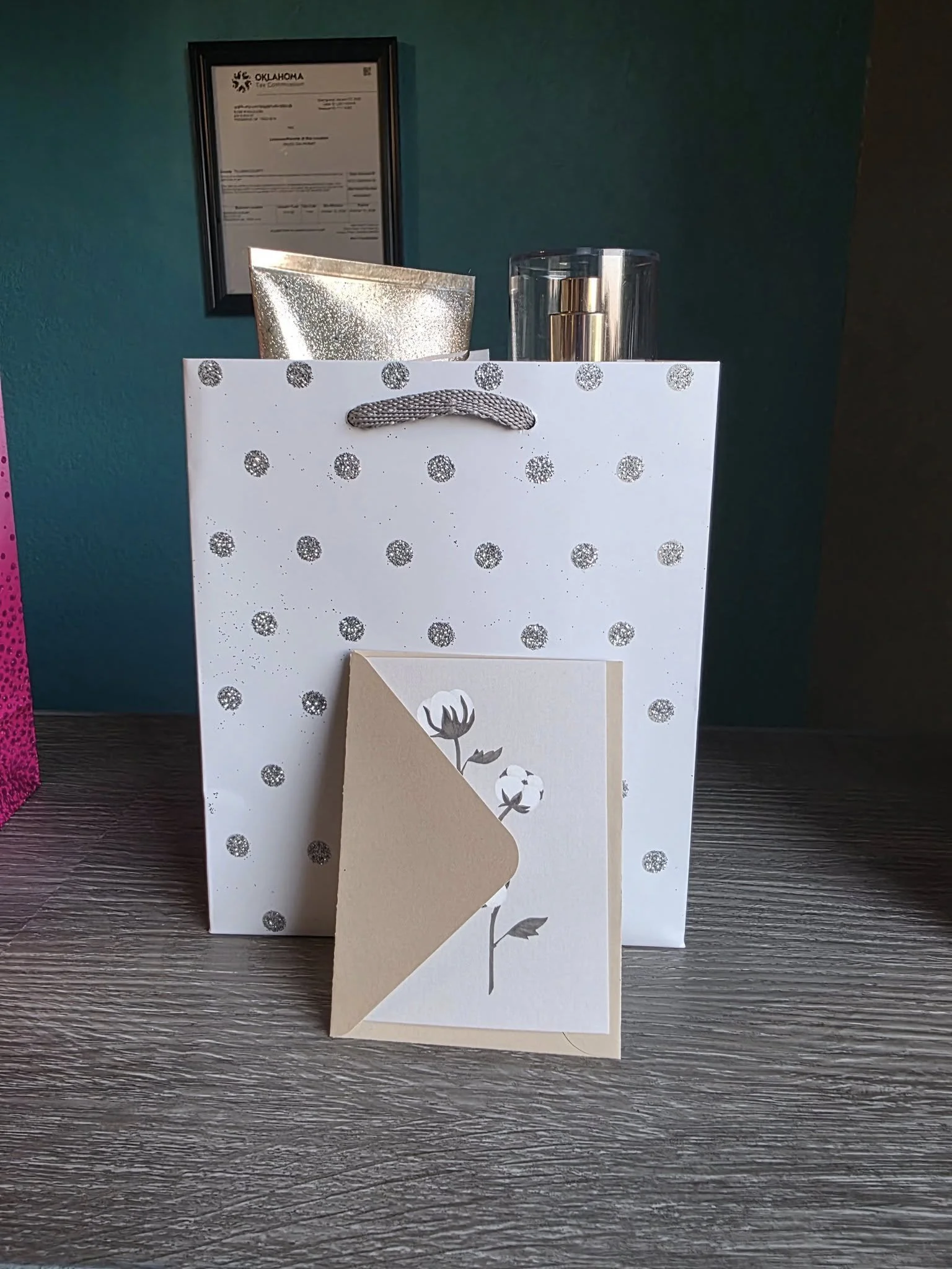 A white gift bag decorated with silver glittery polka dots, placed on a wooden surface. On the bag, there's a greeting card with a floral design. Behind the bag, there is a gold-colored pouch and a clear rectangular container with a metallic gold obj
