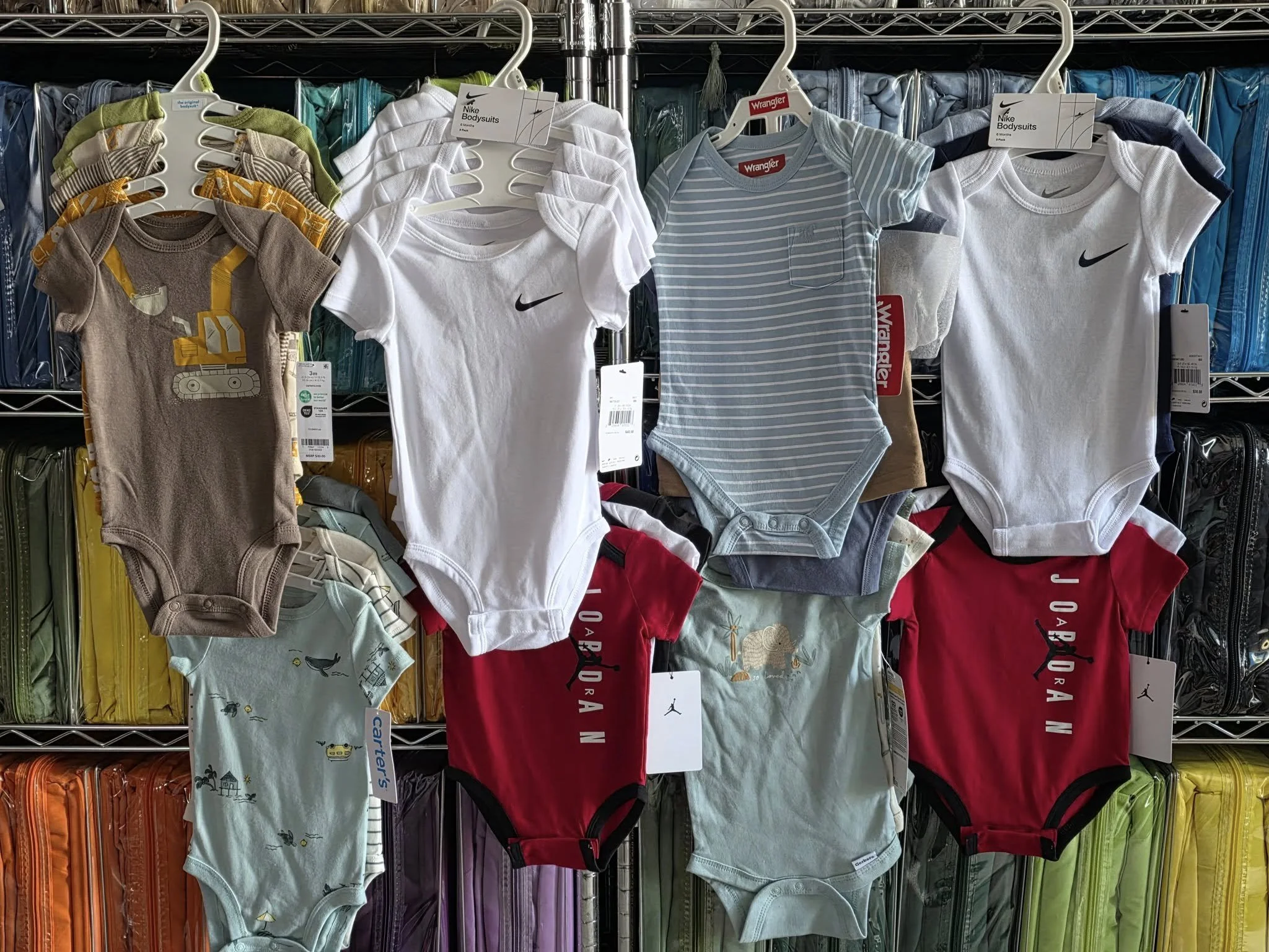 A display of baby onesies hanging on a metal rack, with various colors and patterns, including plain white, striped blue, grey with animal prints, and some with logo designs. Behind the onesies, colorful suitcases are stacked on shelves.