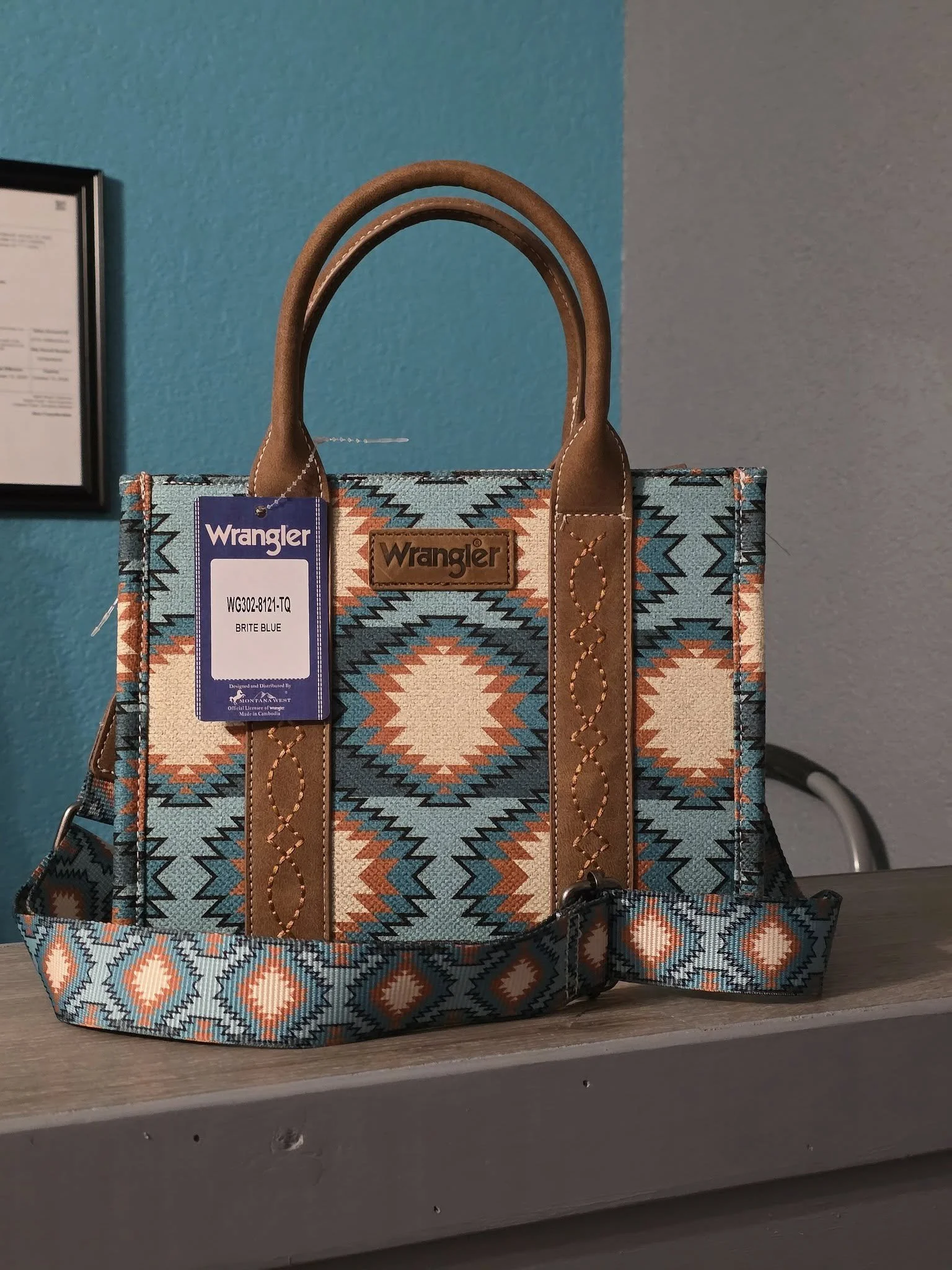 A colorful Wrangler handbag with a southwestern pattern, tan leather accents, and a detachable strap, placed on a gray surface with a blue and gray wall in the background.