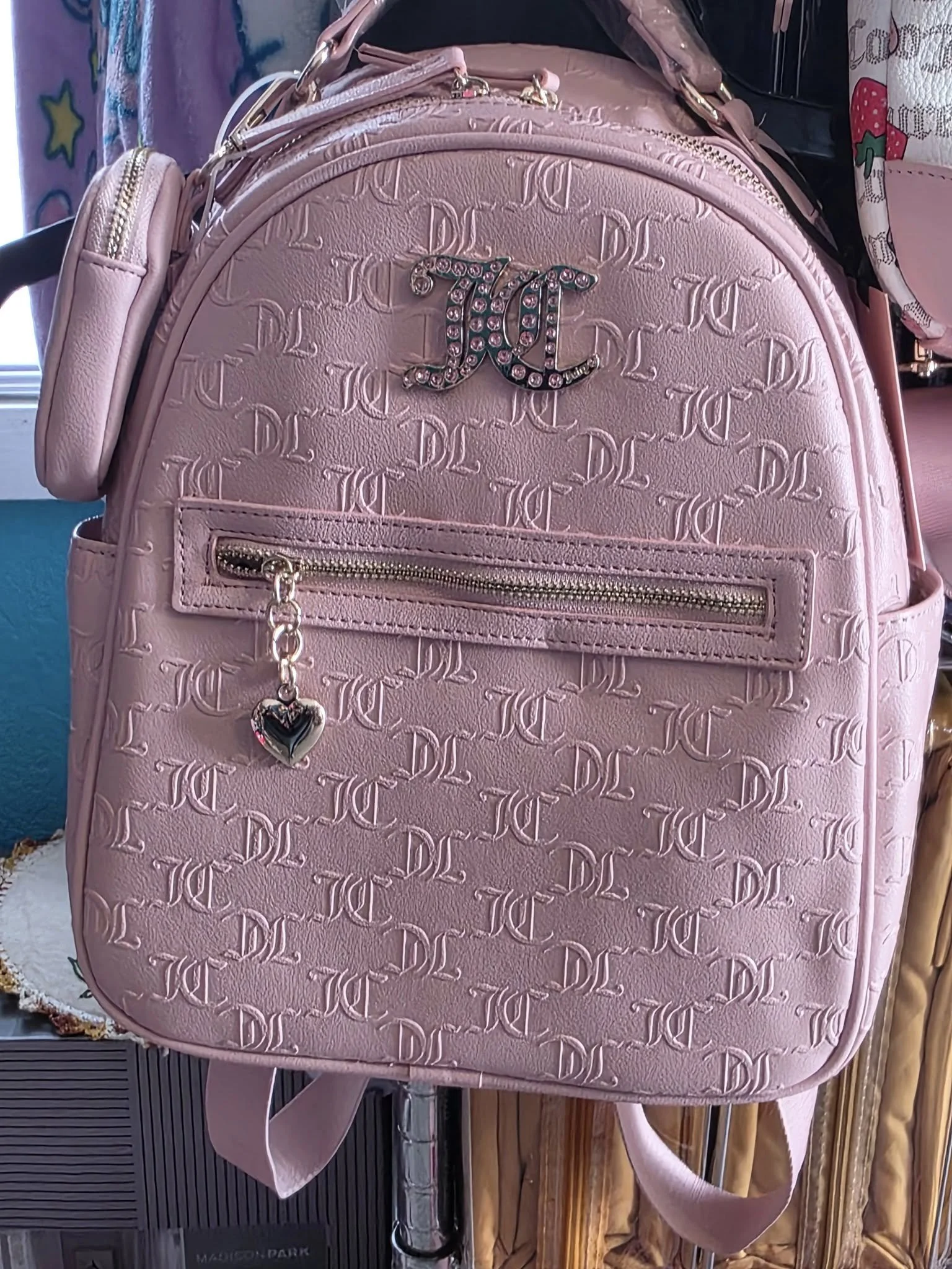 A pink designer backpack with embossed pattern, a metallic logo, and a front zippered pocket with a heart charm.