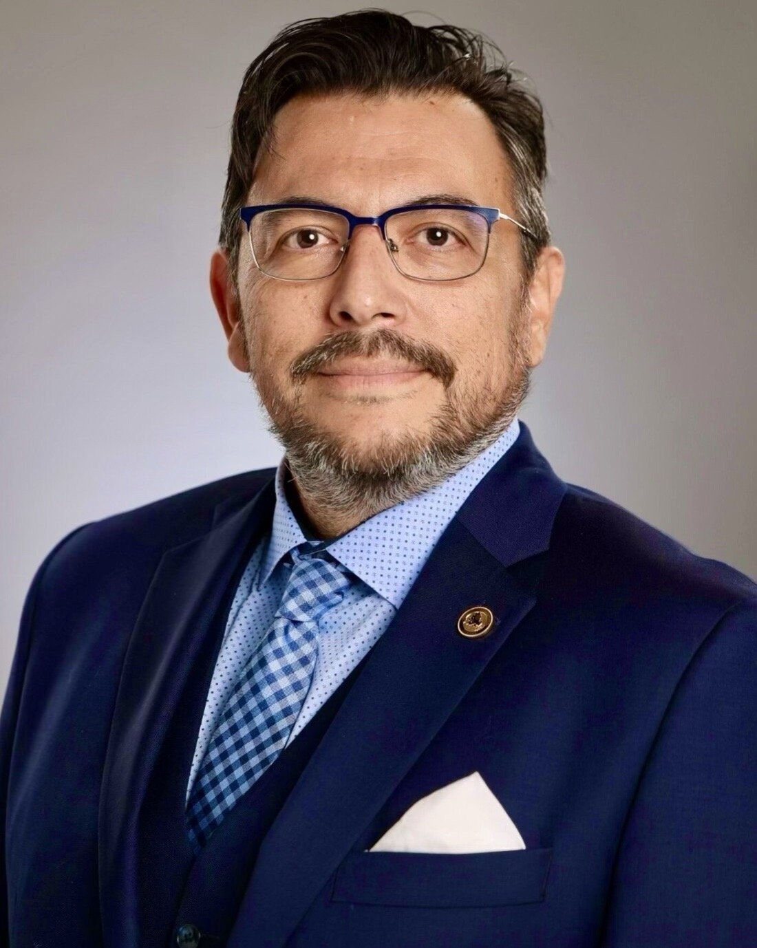 A professional man with glasses, a beard, and a mustache wearing a dark blue suit, light blue shirt with a pattern, a checkered tie, and a white pocket square. He is posing against a plain gray background.