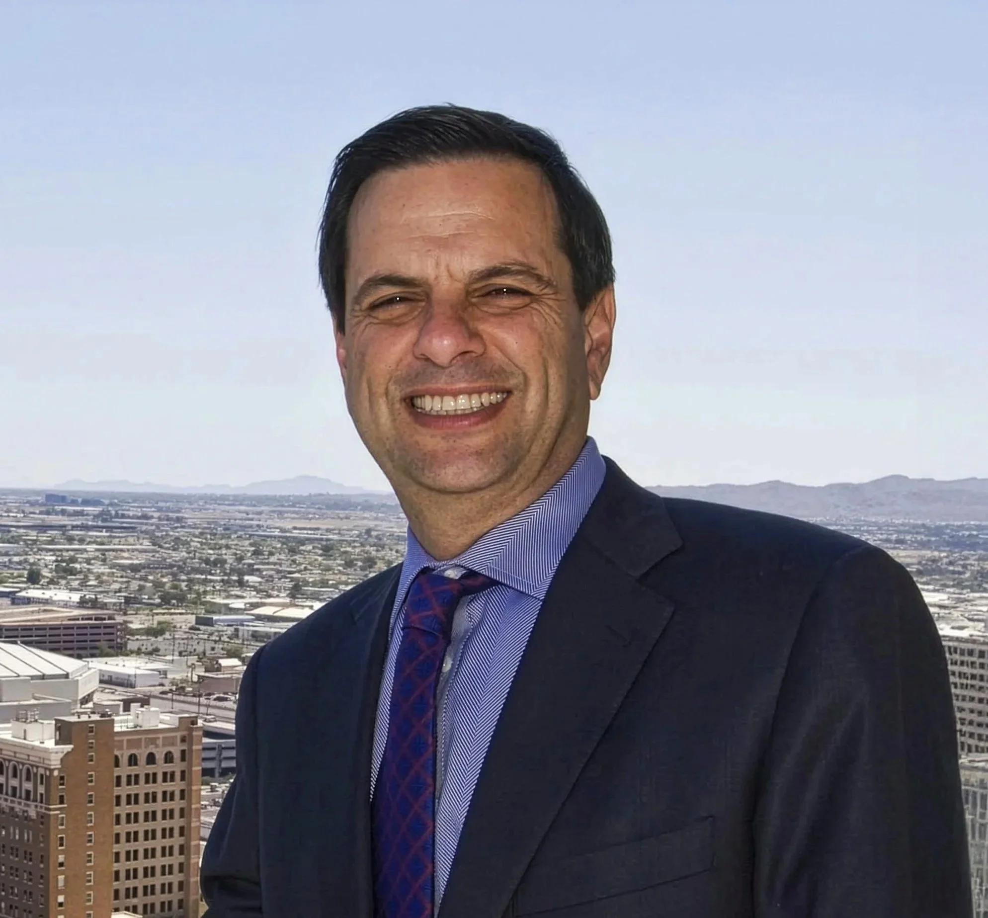 Man in suit smiling on rooftop with cityscape and mountains in background.