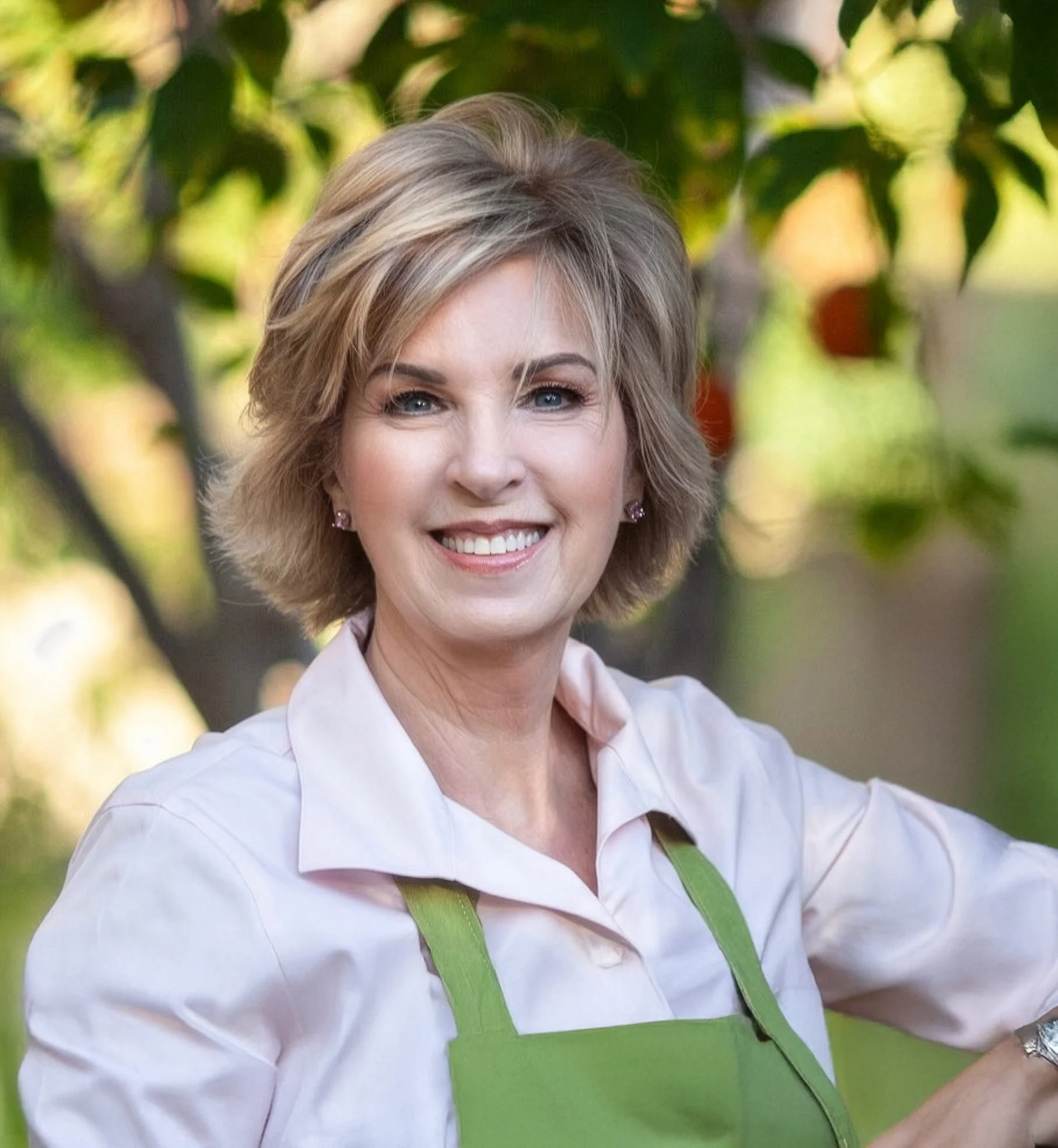 Close-up of a smiling woman with short blonde hair, wearing a peach shirt, with a green apron, standing in front of a blurred background of green leaves.