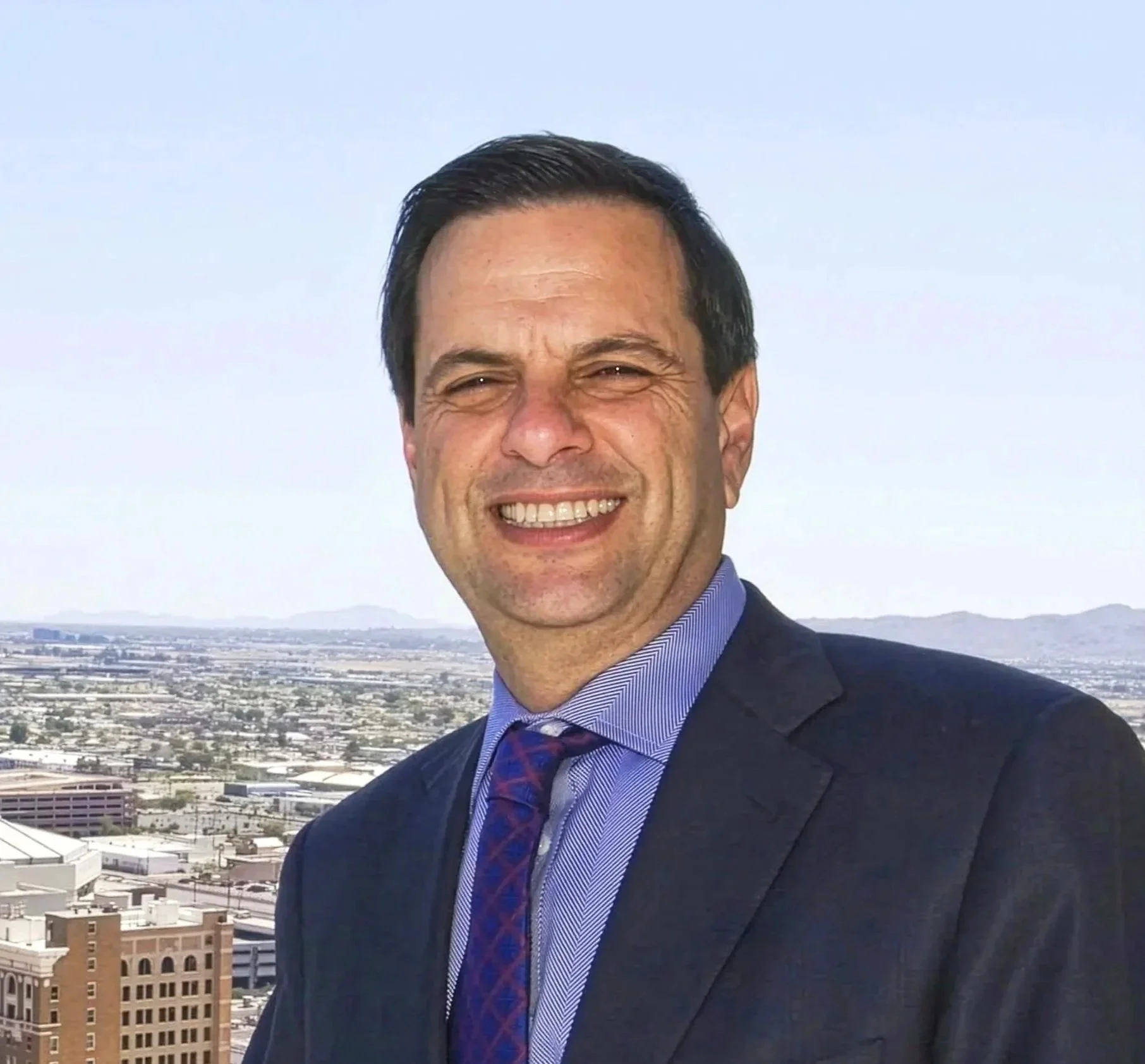 Man in suit smiling on rooftop with cityscape and mountains in background.