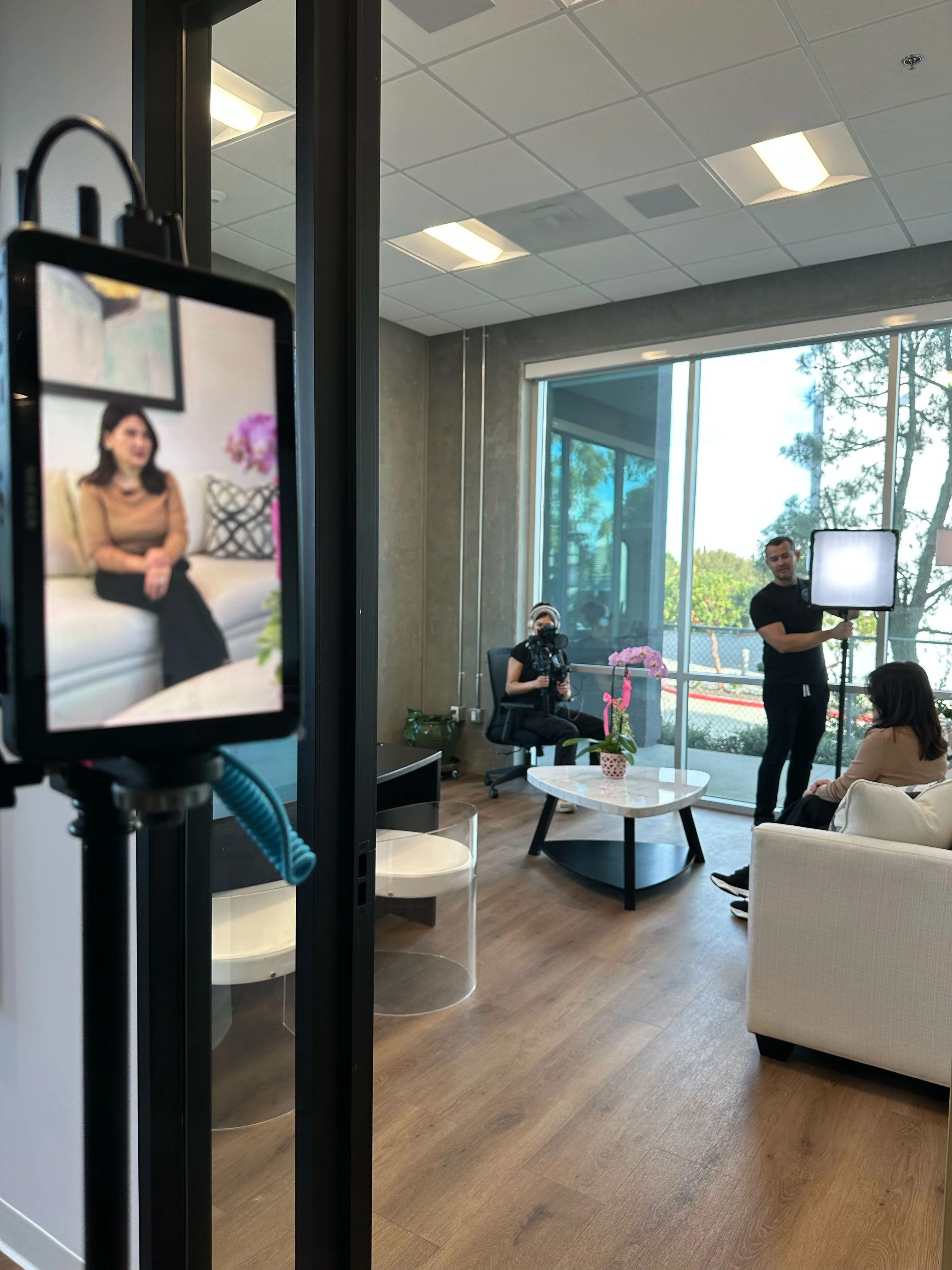 People filming outside a store called Extra Mile with a person dressed in a maroon superhero costume with a yellow and red emblem, in front of a stone and stucco building, under clear blue sky.