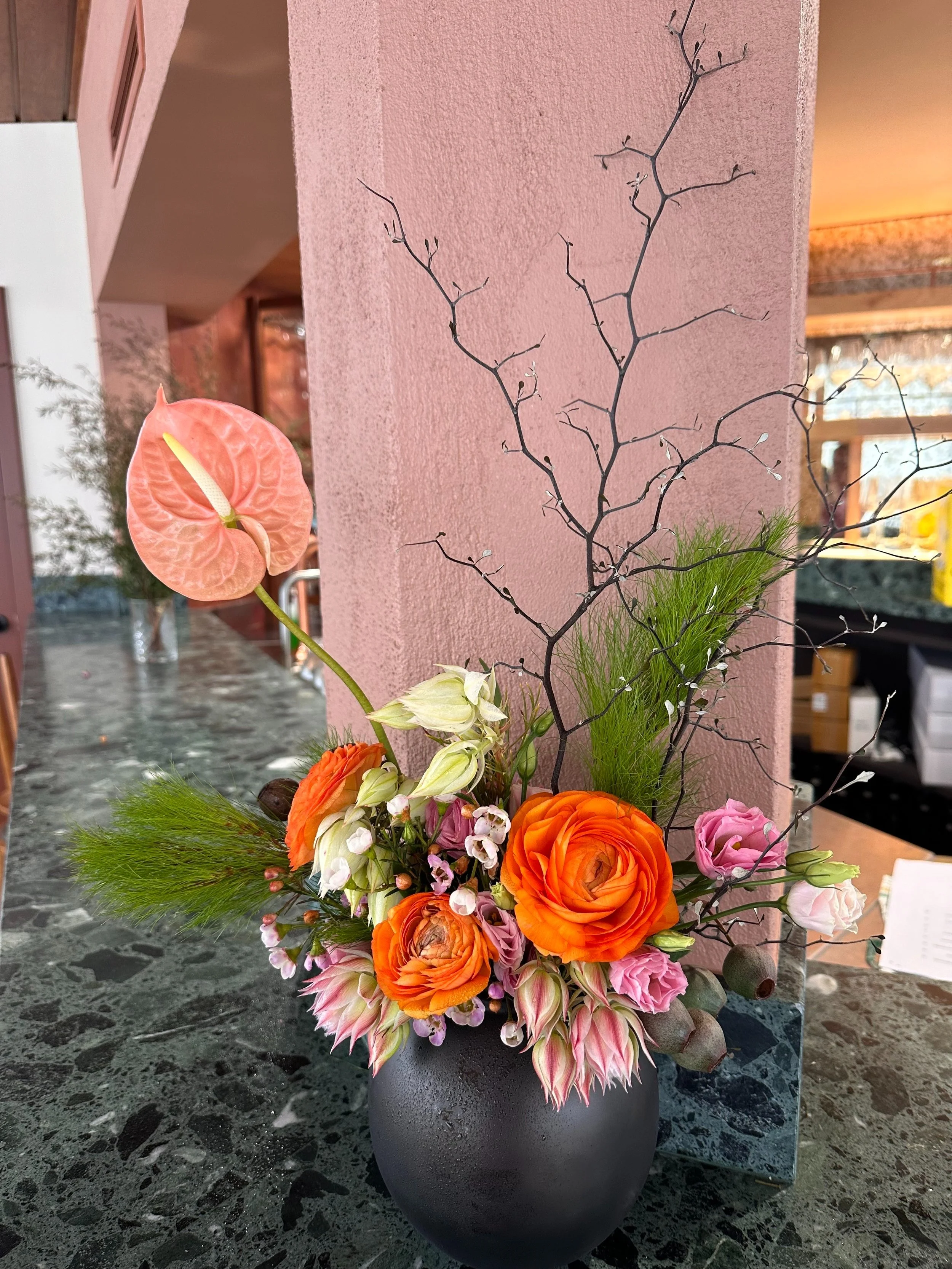A floral arrangement in a black vase with orange, pink, white, and green flowers, including an anthurium, ranunculus, and greenery, placed on a dark speckled countertop against a pink textured wall.