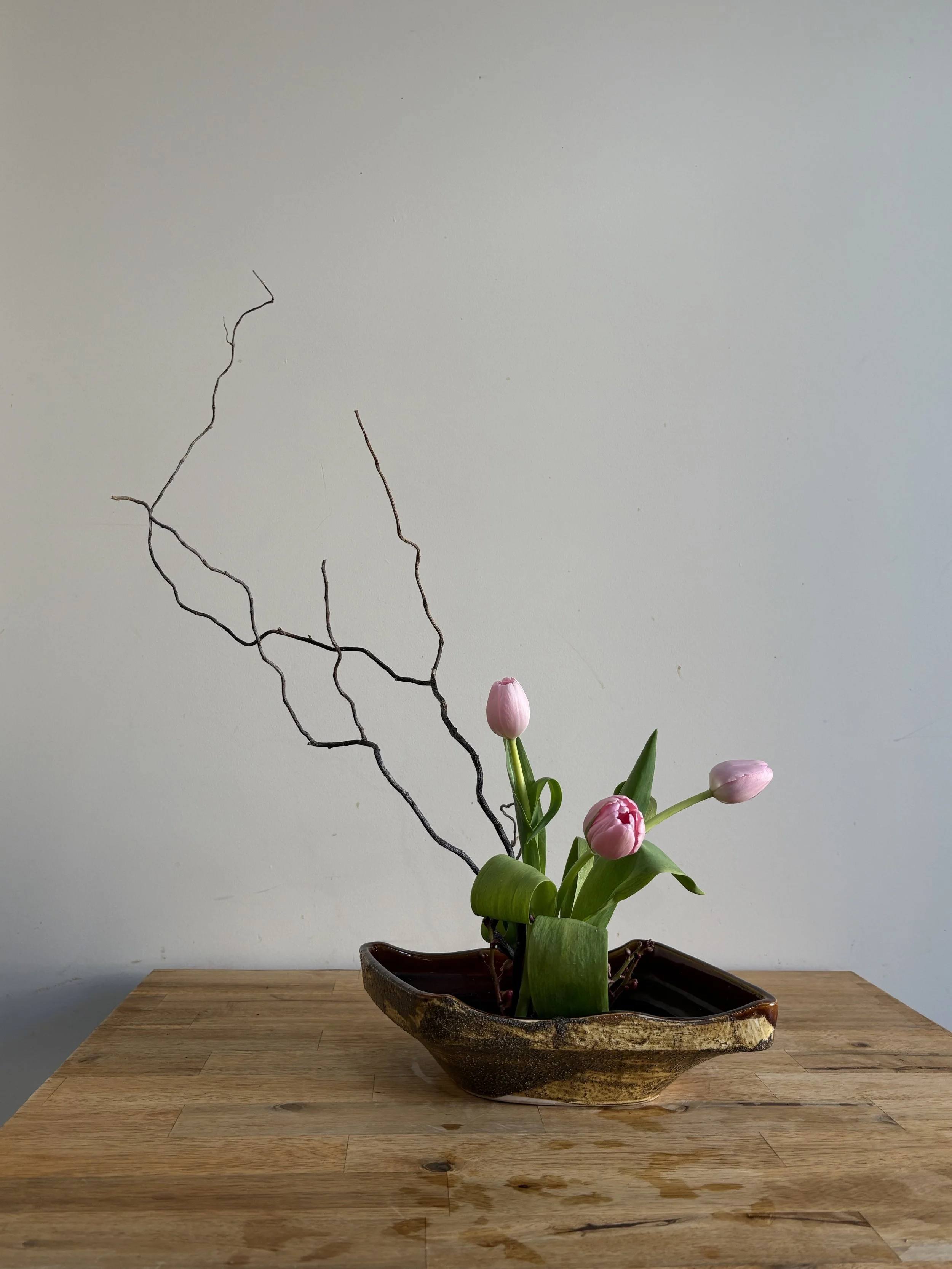 A flower arrangement with pink tulips and twisted branches in a rustic ceramic bowl on a wooden table.