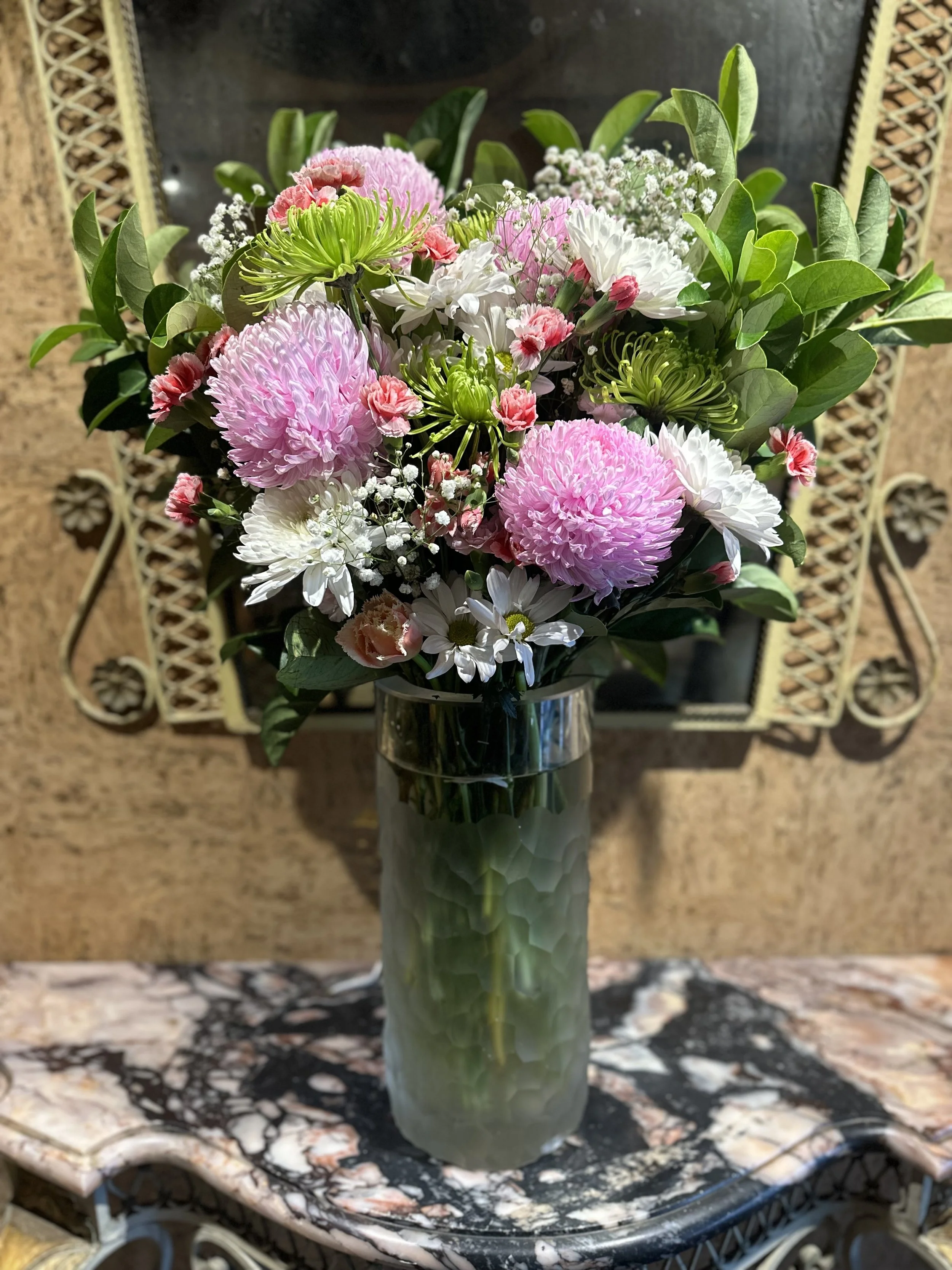 A bouquet of pink, white, and green flowers in a tall, textured green glass vase, placed on a marble-top table with a decorative wrought iron frame, in front of a mirror with a gold ornate frame.