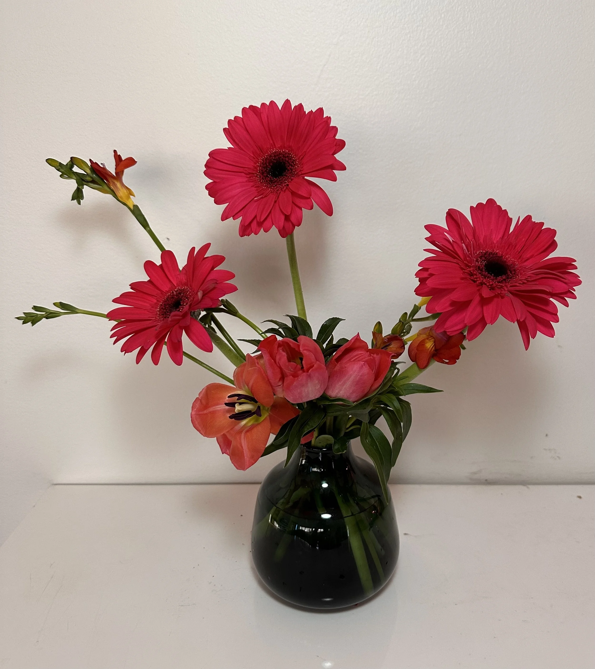 A black vase holding pink and red flowers, including gerbera daisies, alstroemeria, and buds, set against a white wall.