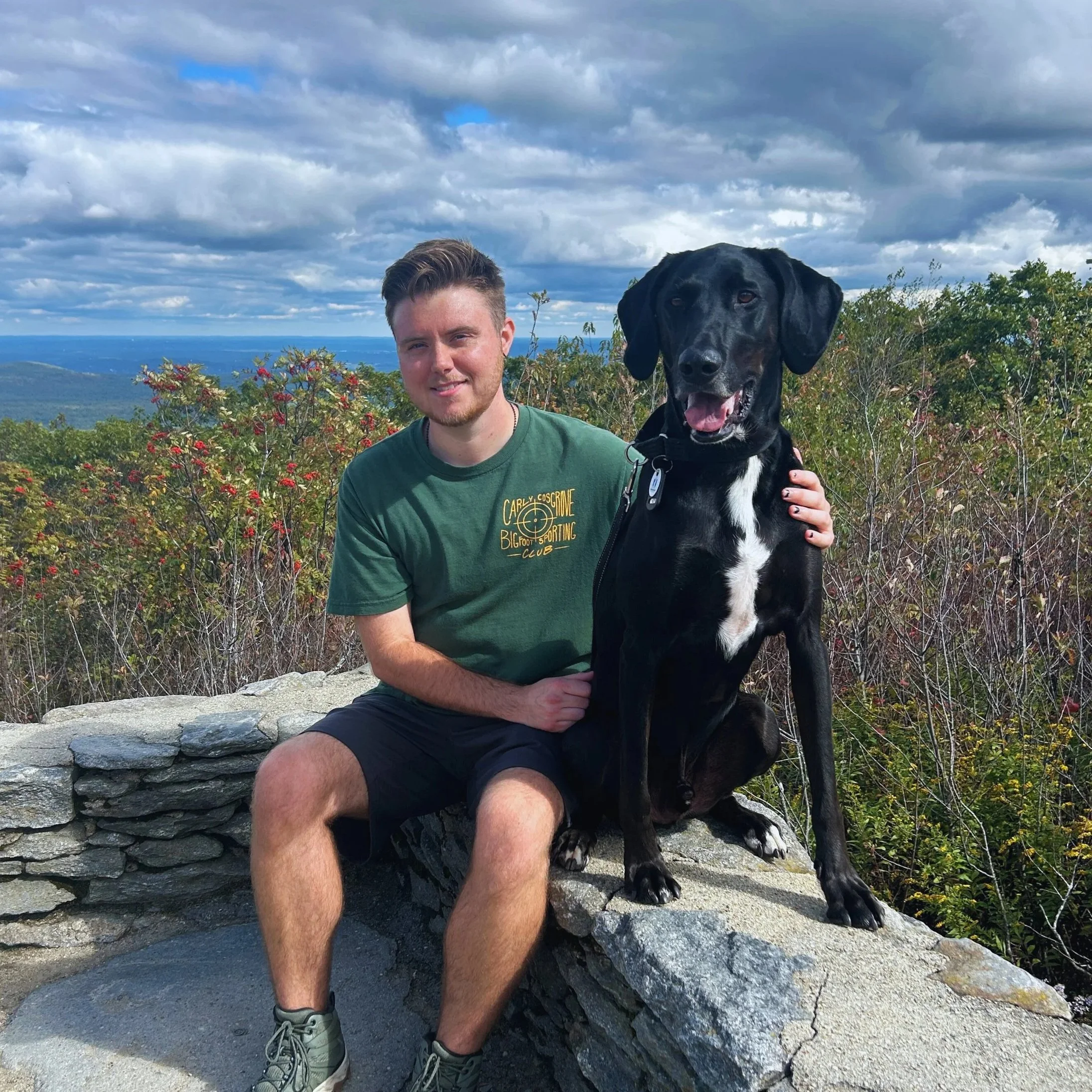 A young man sitting on a stone wall outdoors with his black dog, both smiling, with trees and mountains in the background under a partly cloudy sky.