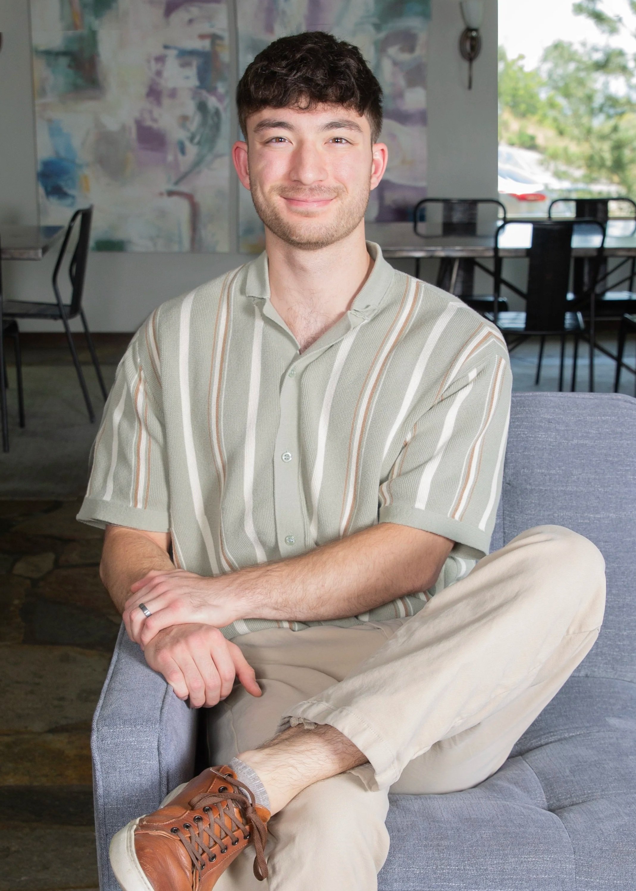 Ira Bennett, Milieu Manager at Eva Carlston Academy, sitting comfortably on a gray sofa with a warm smile, wearing a short-sleeved green striped shirt and khaki pants.