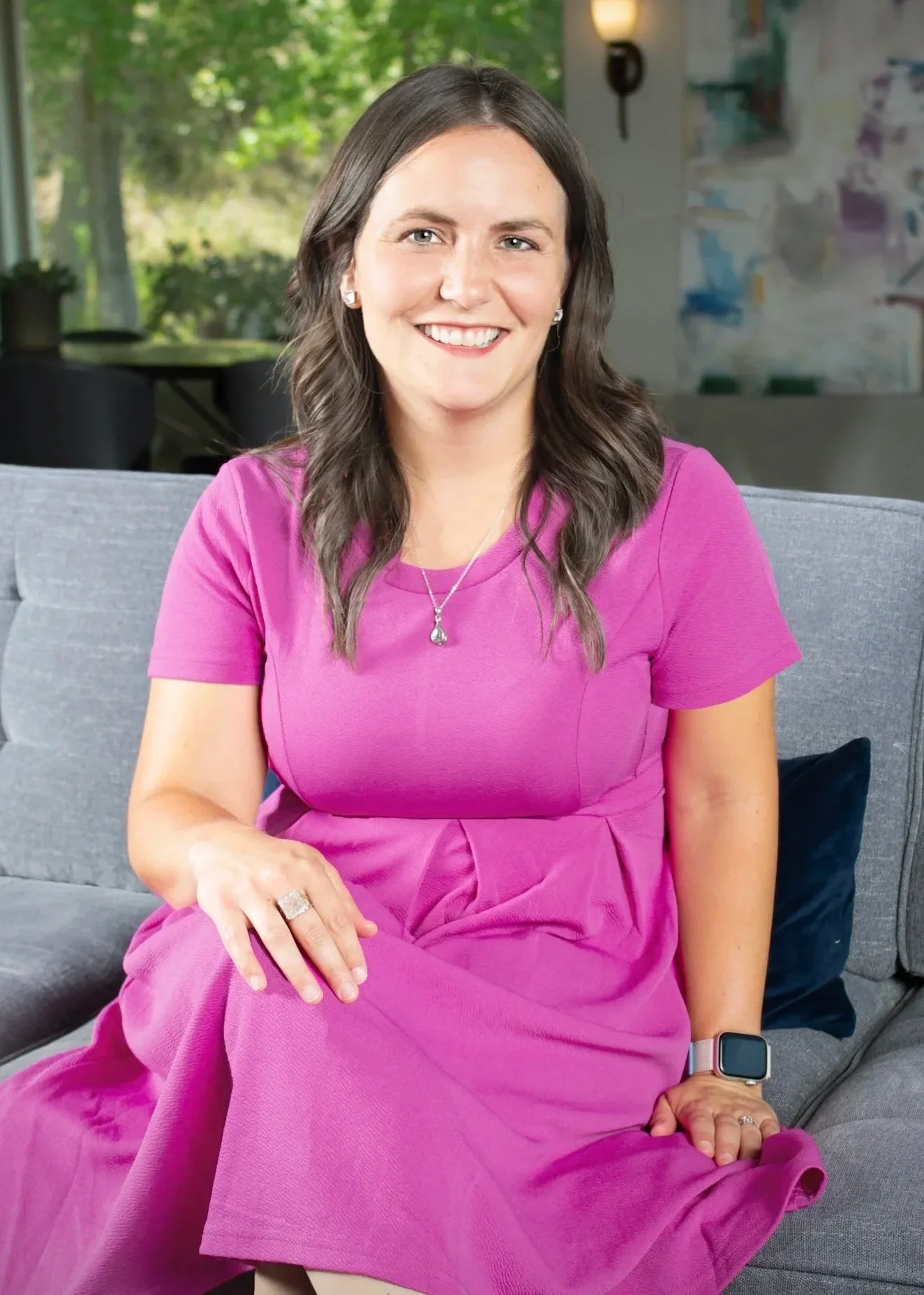 Smiling woman with shoulder-length dark hair wearing a bright pink dress, seated on a gray couch indoors.