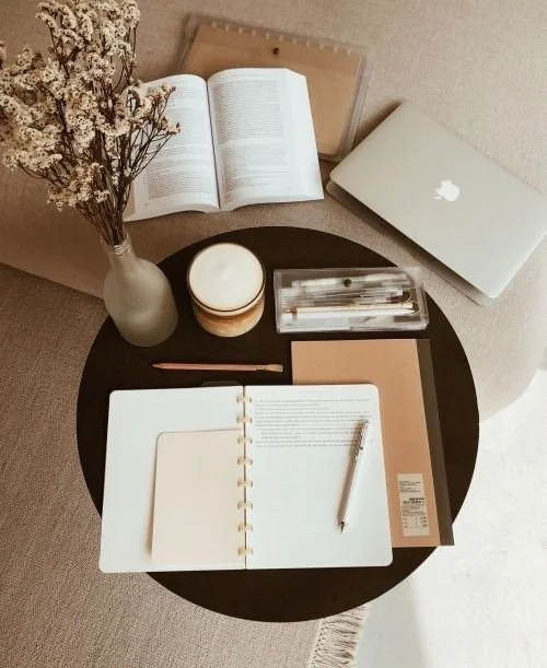 A round black table with a vase of dried flowers, an open planner, a candle, a small box, a closed notebook, a pen, an open book, and a closed MacBook laptop.