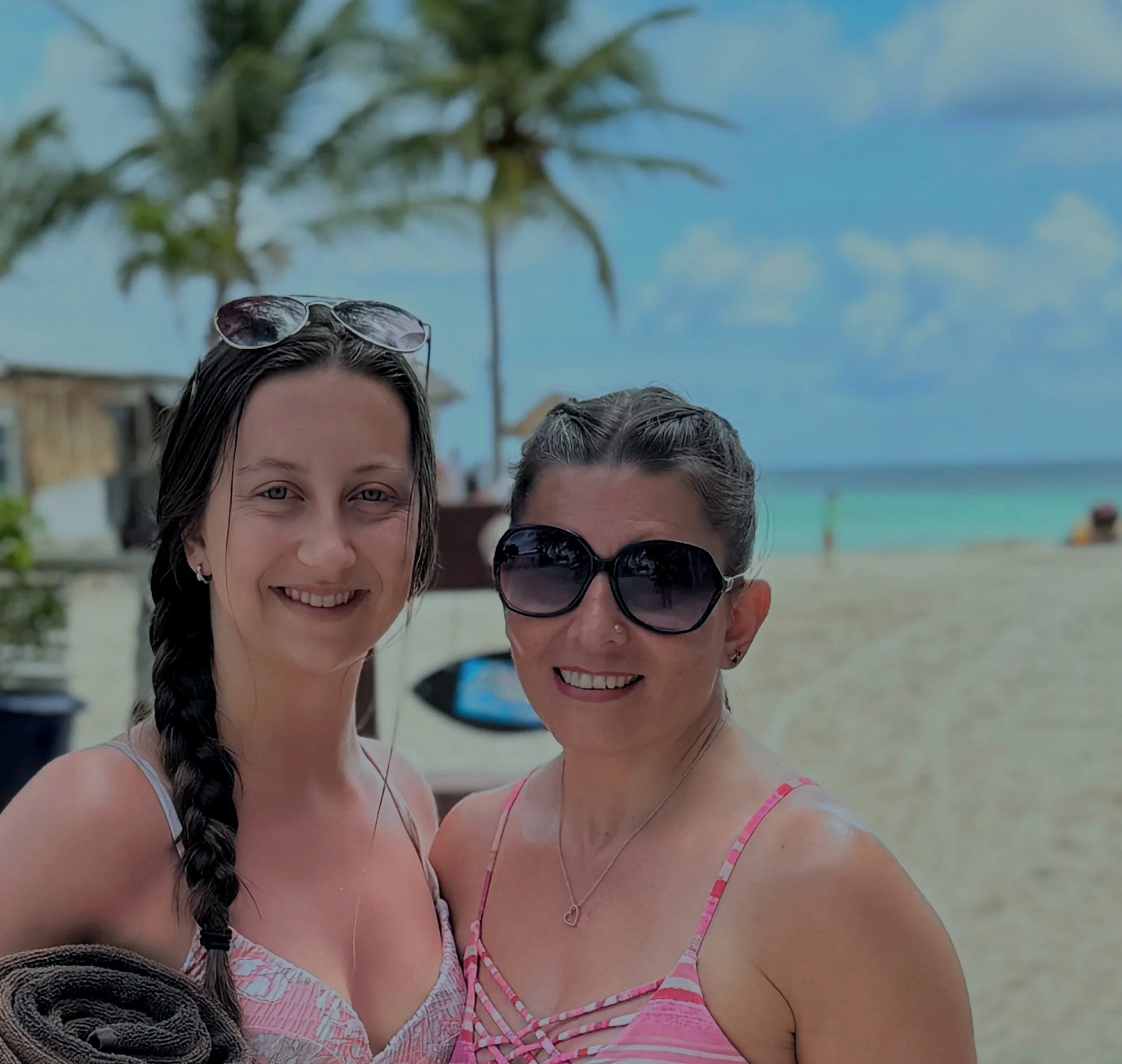 Two women at the beach, one with long braided hair wearing a pink and white striped dress, and one with short hair wearing sunglasses. There are palm trees and the ocean in the background.