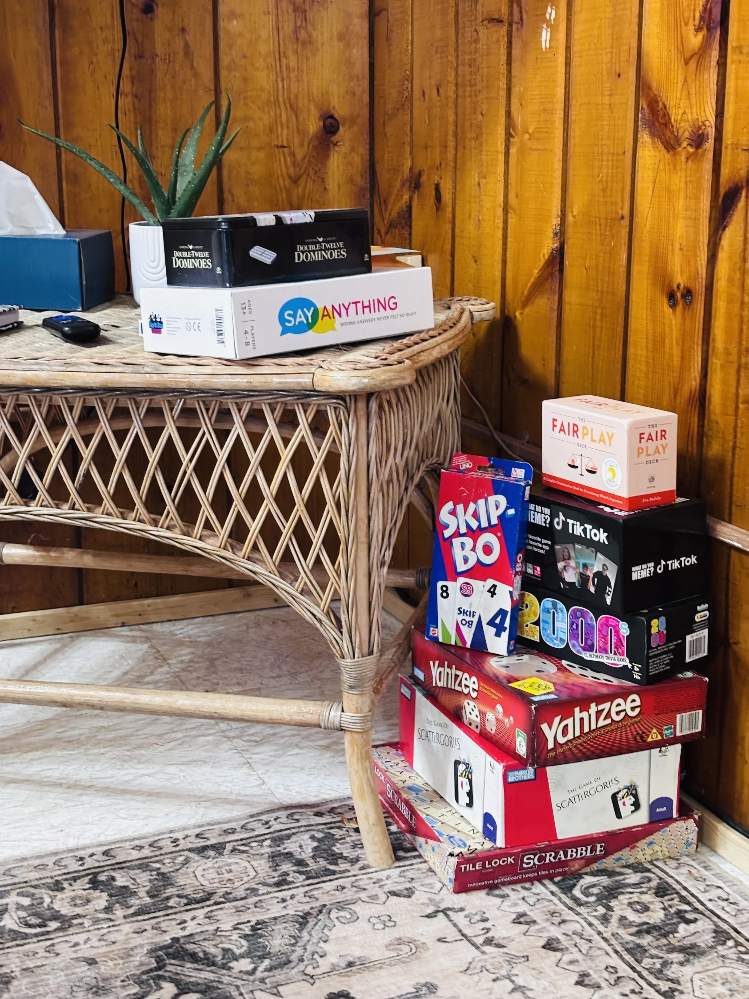 Stacked board games including Yahtzee, Skip-Bo, Tetris, Scattergories, and Tile Lock Scrabble in a room with wooden walls and a wicker table.