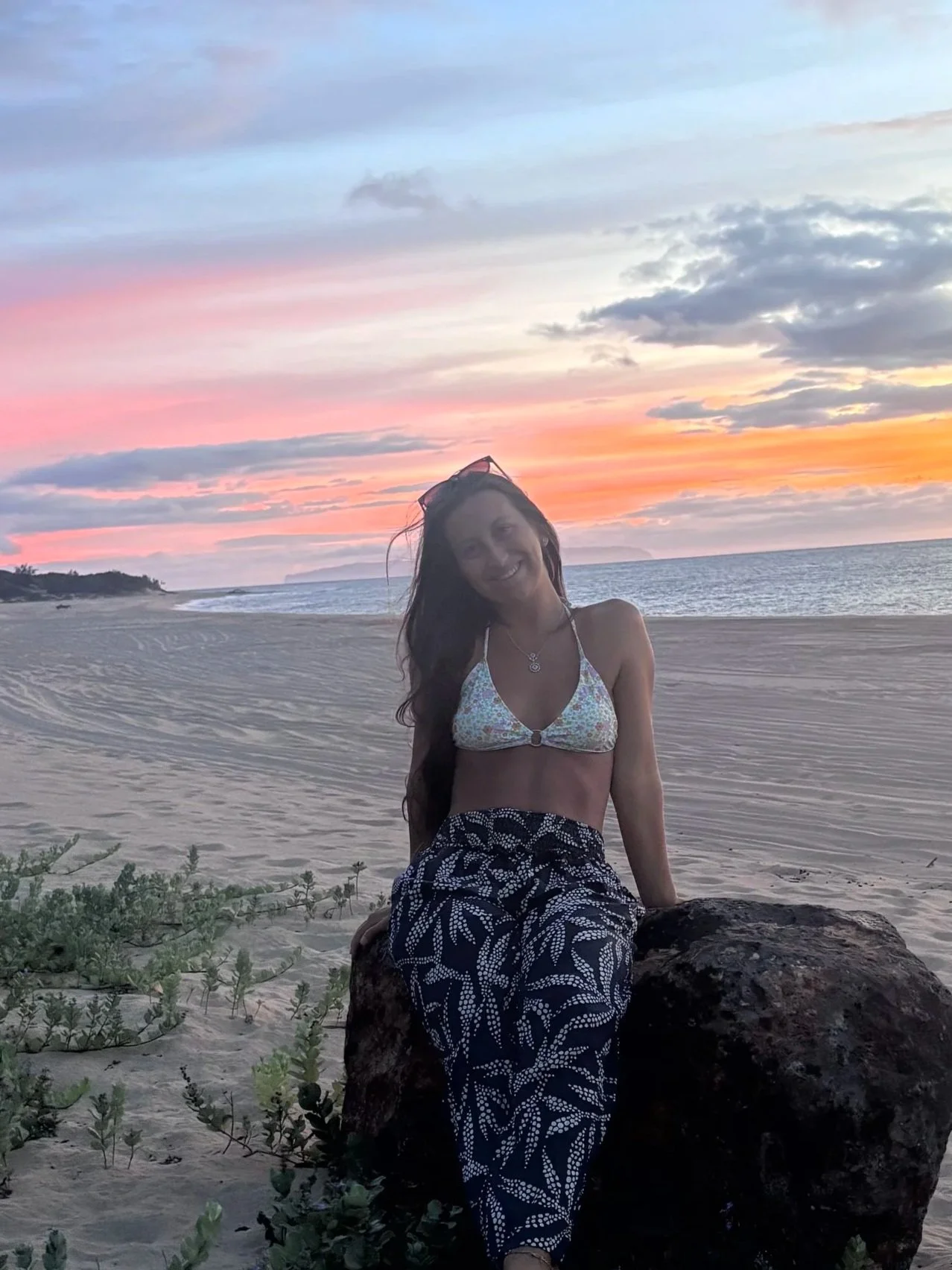 A woman sitting on a rock on a sandy beach at sunset, smiling, with ocean waves and a colorful sky in the background.