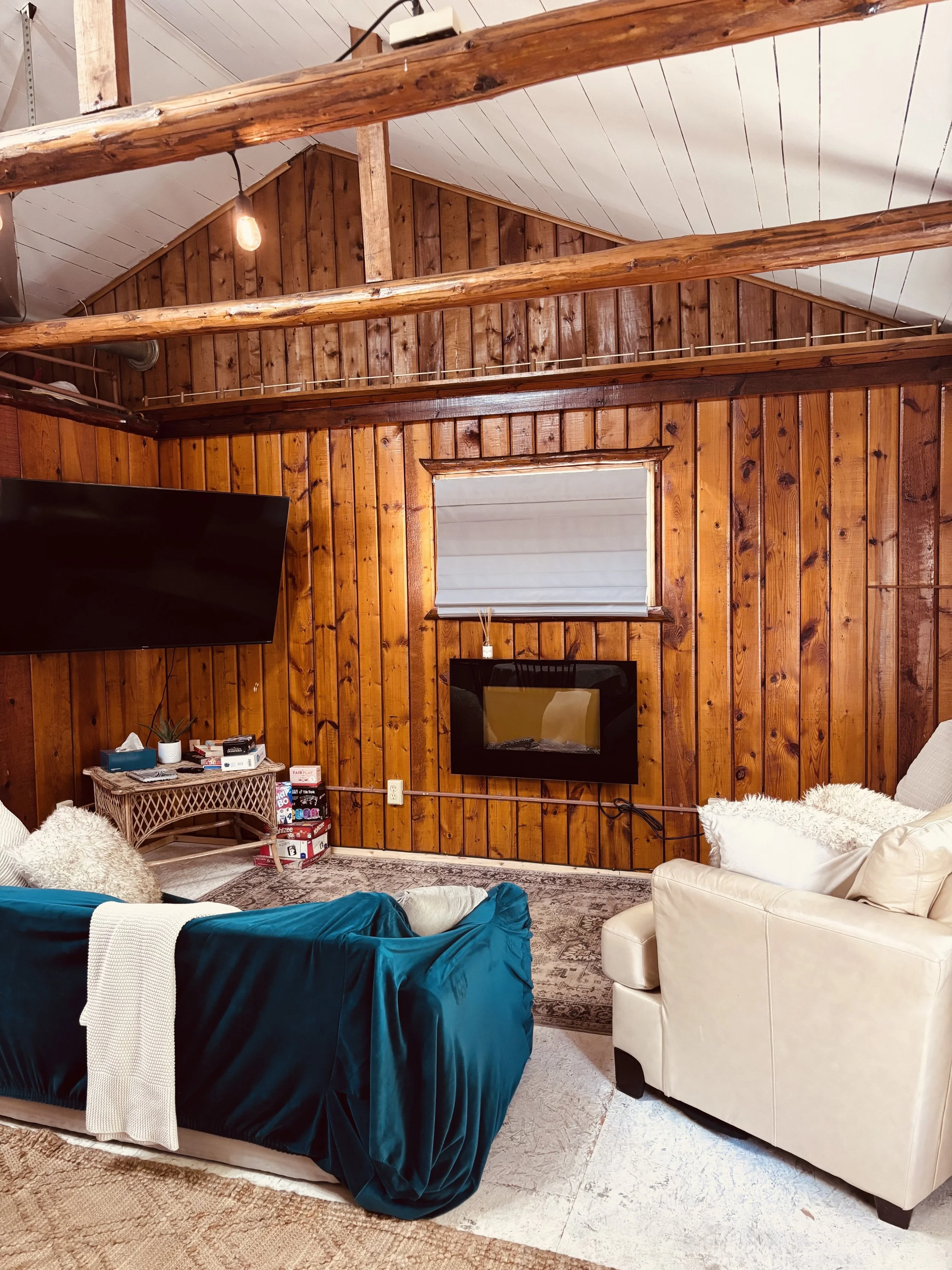 Living room with wooden panel walls, a white armchair, a teal couch with a white blanket, wall-mounted TV, small side table with decor, window with blinds, carpeted floor, and exposed ceiling beams.
