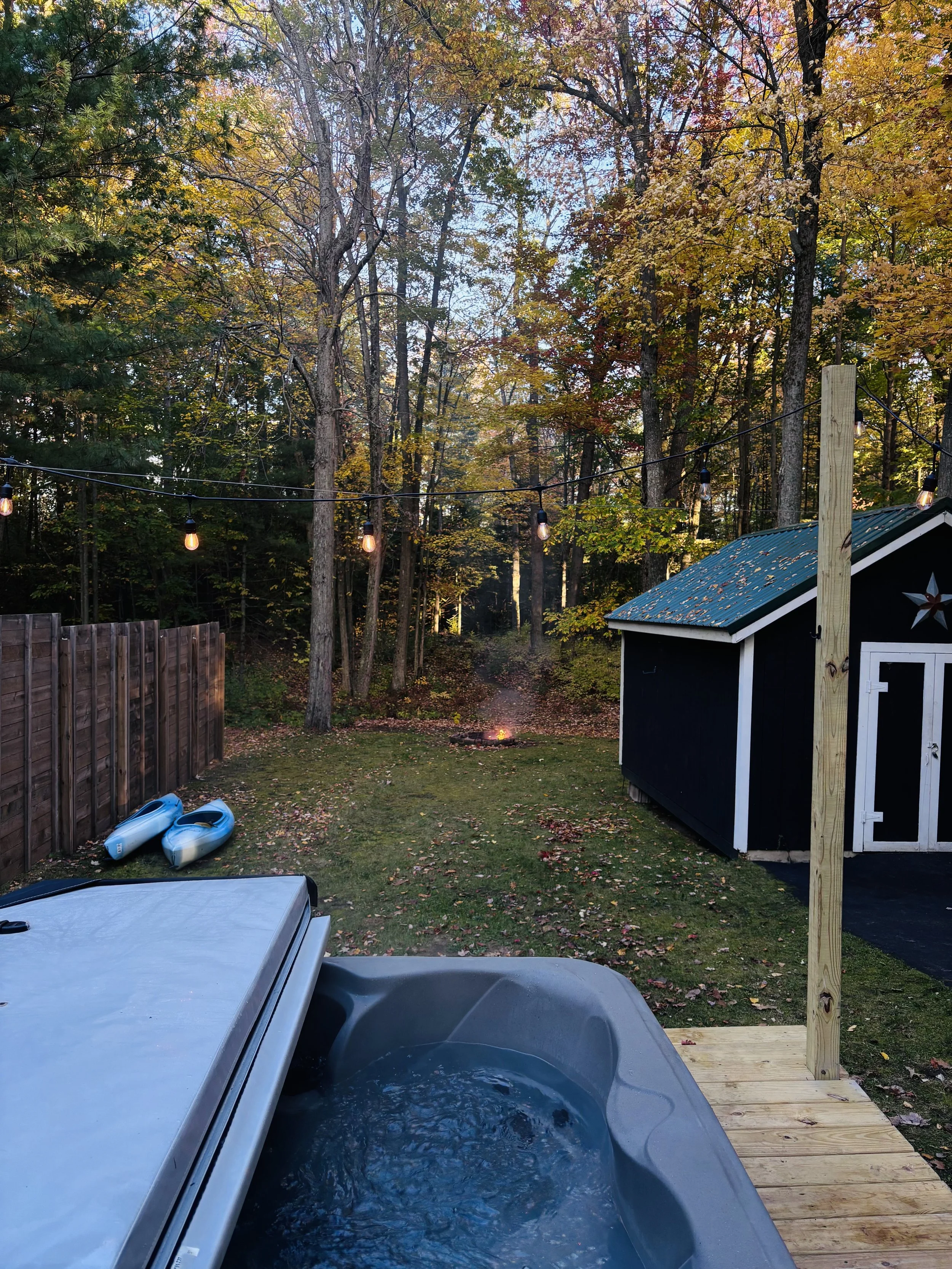 Backyard with hot tub, string lights, wooden fence, kayak, shed, fire pit, trees with autumn leaves, and a small wooden platform.