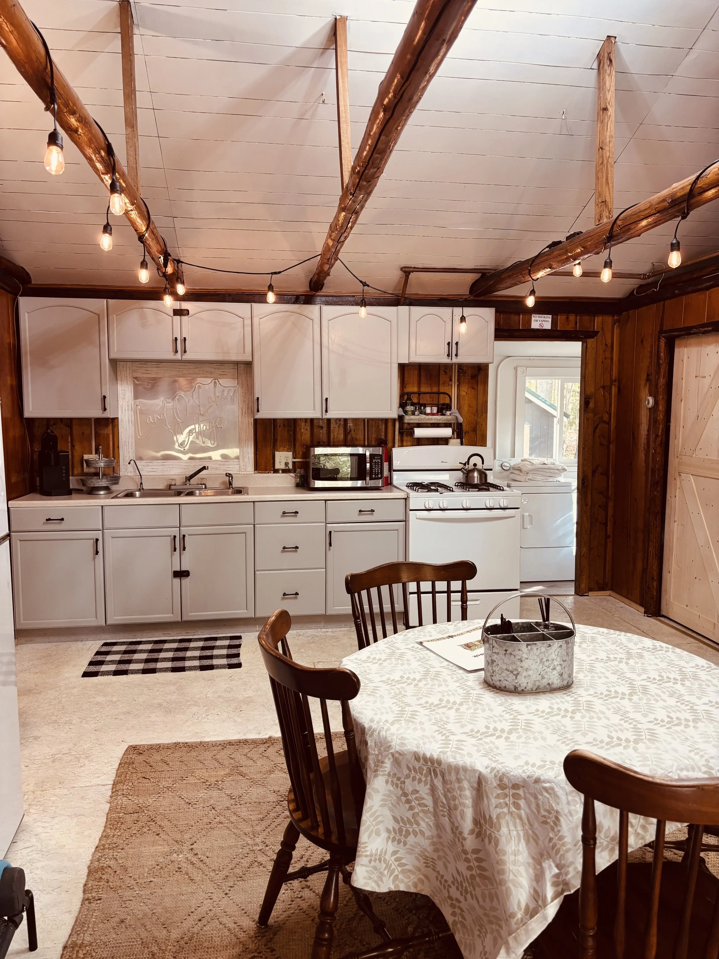 Cozy rustic kitchen with white cabinets, wooden walls, exposed ceiling beams with string lights, a round dining table with a floral tablecloth and chairs, small checkered mat by sink, and a white stove and microwave.
