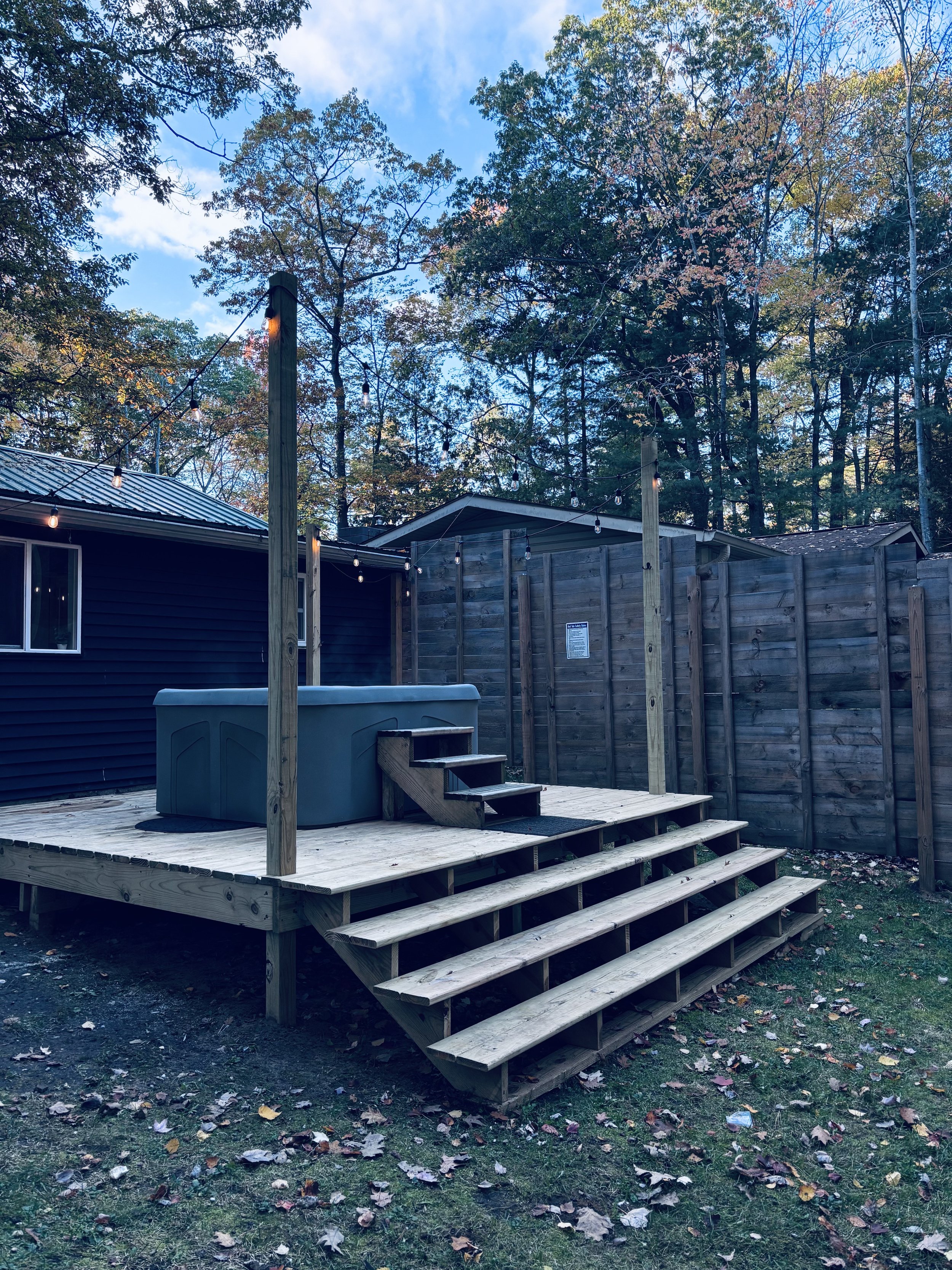 Backyard with a small wooden deck, a hot tub, string lights, surrounded by wooden fencing and trees with fall foliage, under a partly cloudy sky.