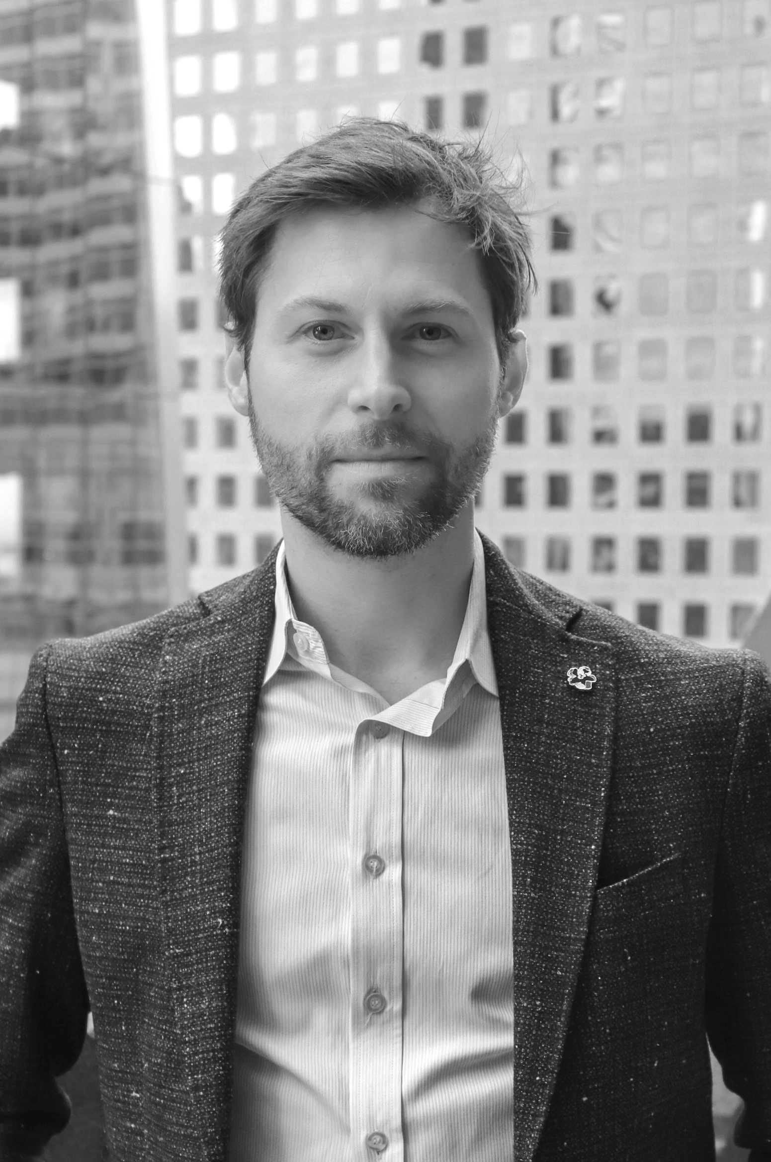 Black and white portrait of a man with glasses, beard, and mustache, wearing a black V-neck T-shirt and dark pants, standing with hands behind back, in a studio with plain background.