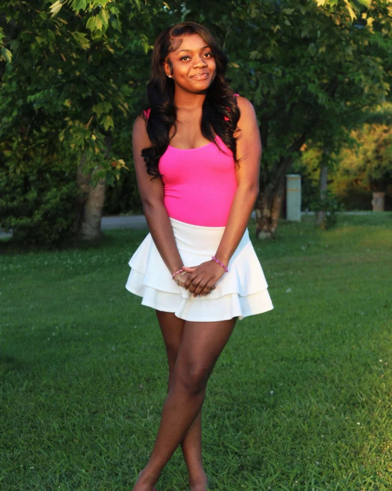 Young woman standing outdoors on grass with trees in the background, wearing a pink top and white skirt, smiling softly.