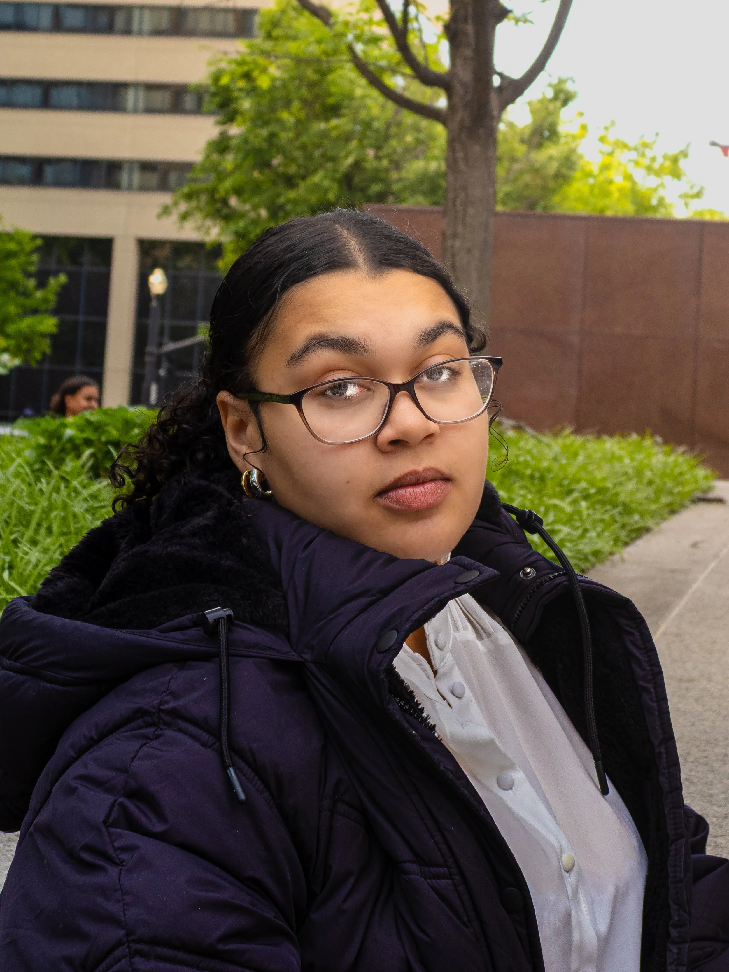 Close-up of a woman wearing glasses, a white shirt, and a dark jacket, outdoors in an urban area with greenery and buildings in the background.