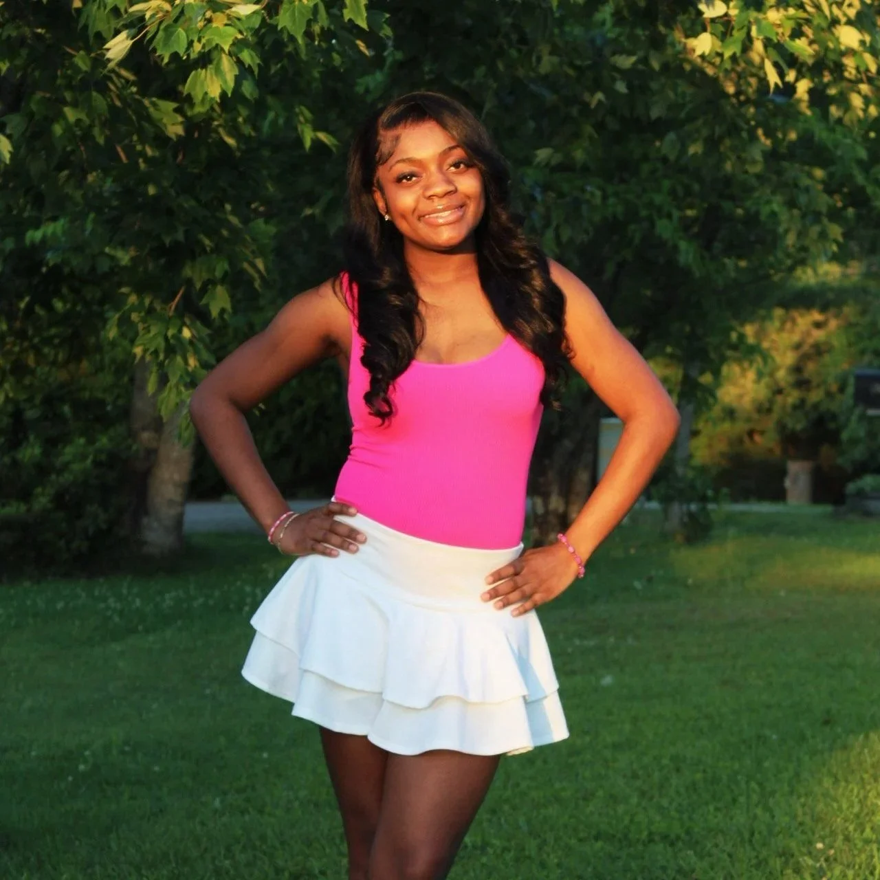 Young woman smiling outdoors in a pink top and white ruffled skirt with her hands on her hips, standing on grass with trees in the background.