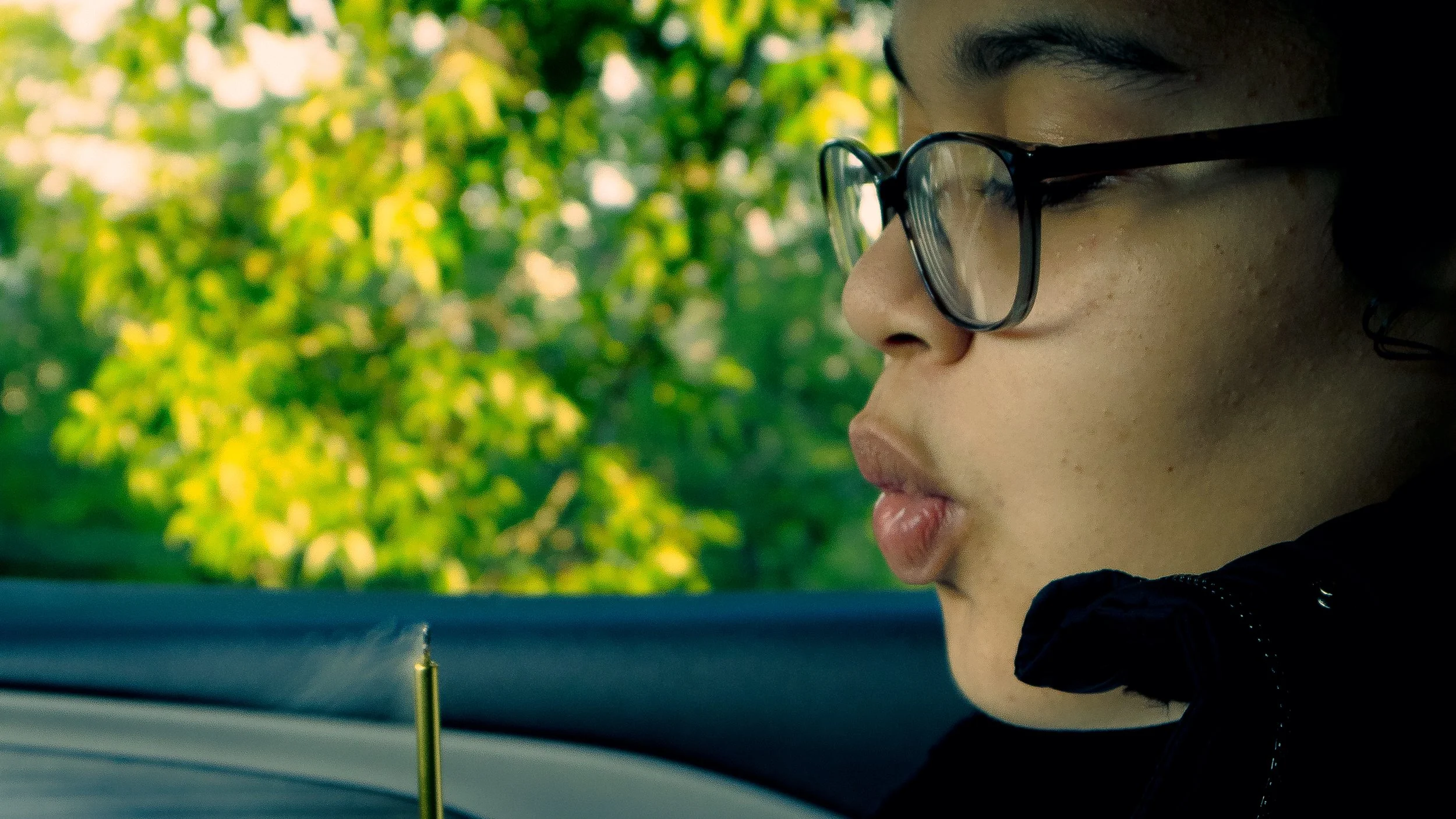Close-up side profile of a young person wearing glasses, focused on an activity, with a blurred background of green foliage.