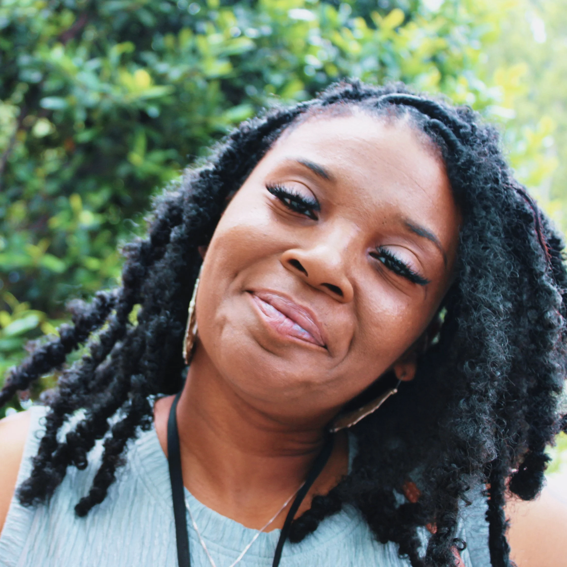Close-up of a smiling woman's face with dark curly hair, outdoors with green foliage in the background.