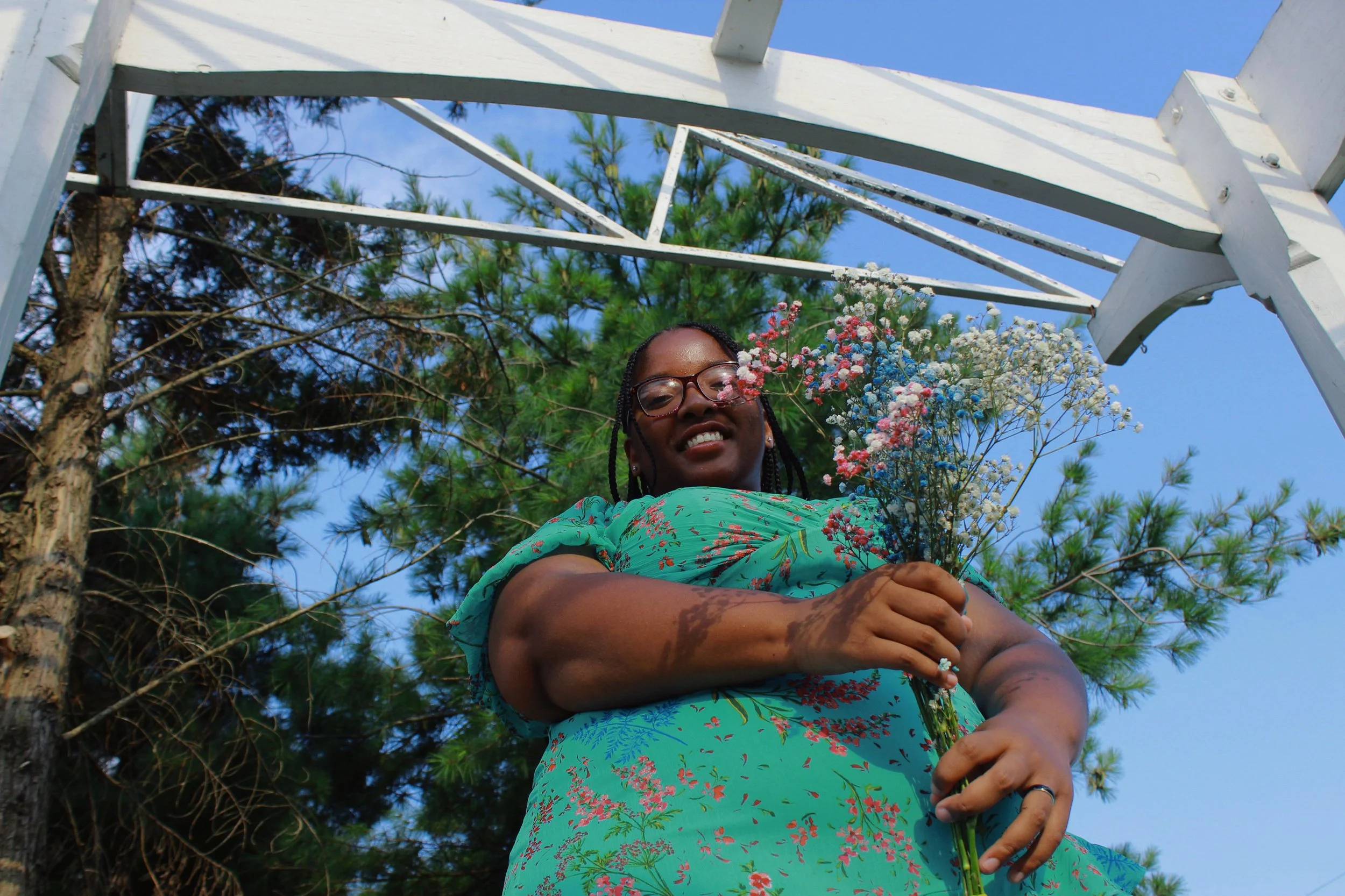 A woman with braids, glasses, and earrings holding a bouquet of colorful flowers, smiling, outdoors under a white wooden structure with trees and a blue sky in the background.