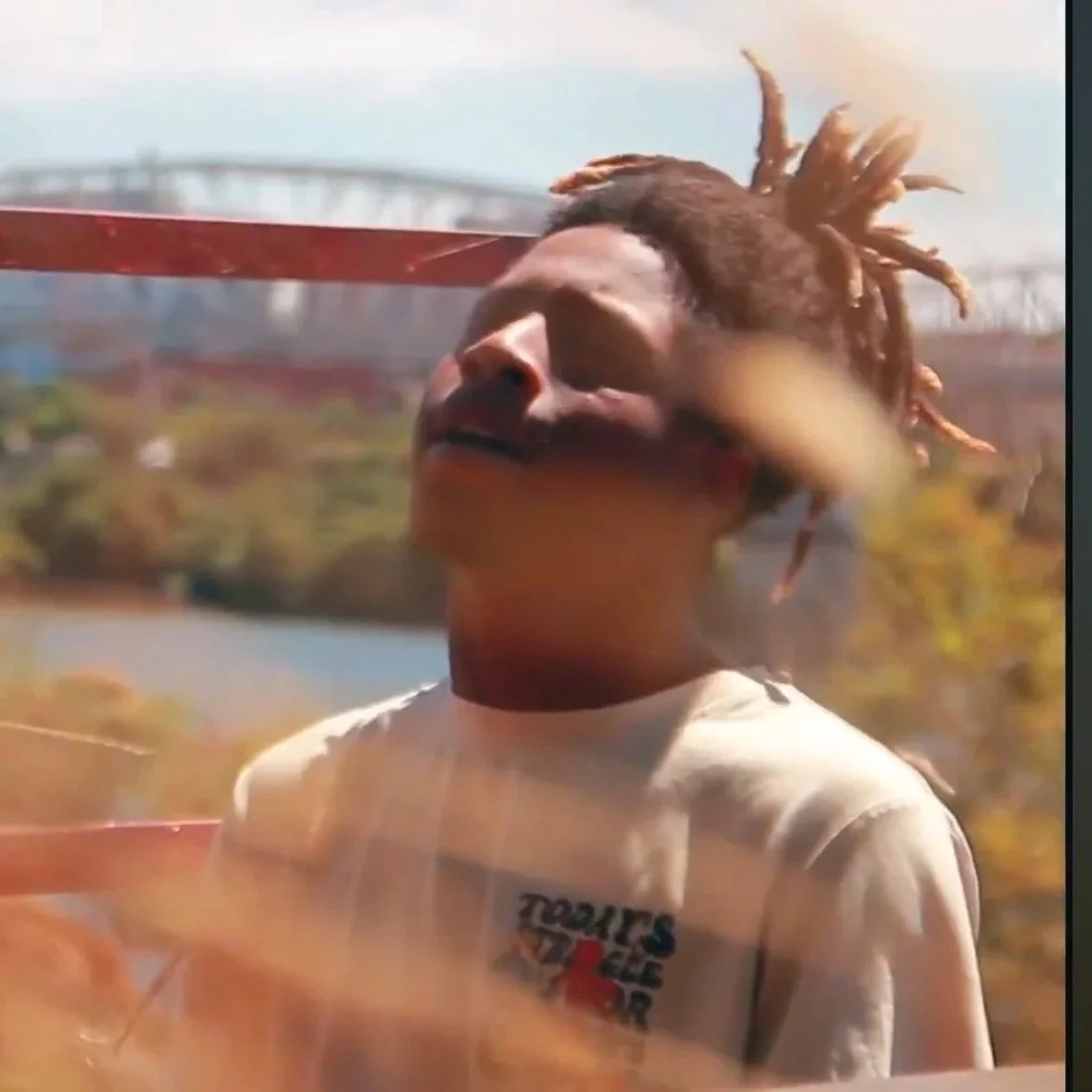 Person with dreadlocks and a white T-shirt standing outdoors, with a blurred background of trees and a bridge.