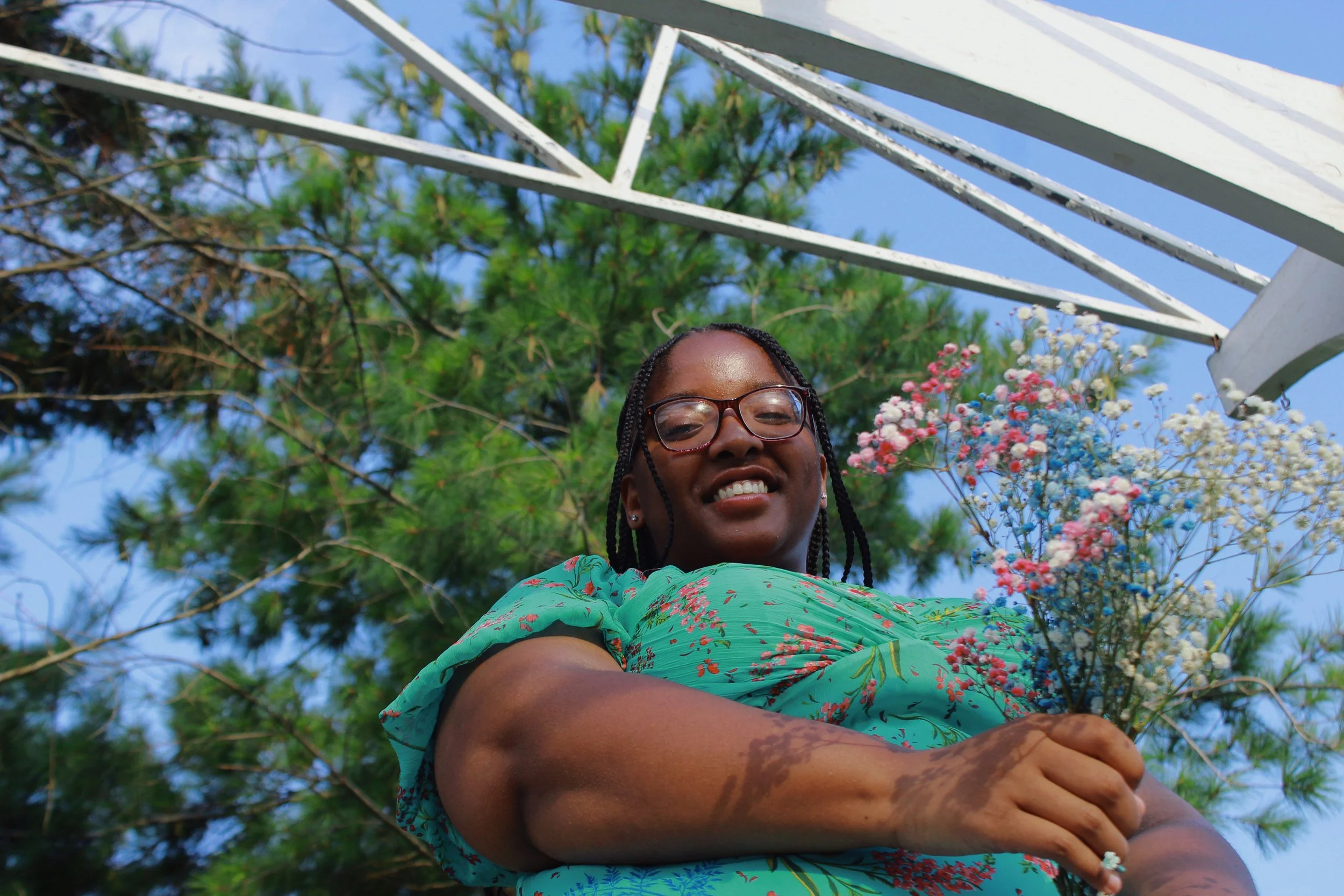 A woman with braided hair and glasses is smiling at the camera, standing outdoors beneath a white metal structure with green trees and blue sky in the background. She is wearing a green floral dress and holding colorful flowers.