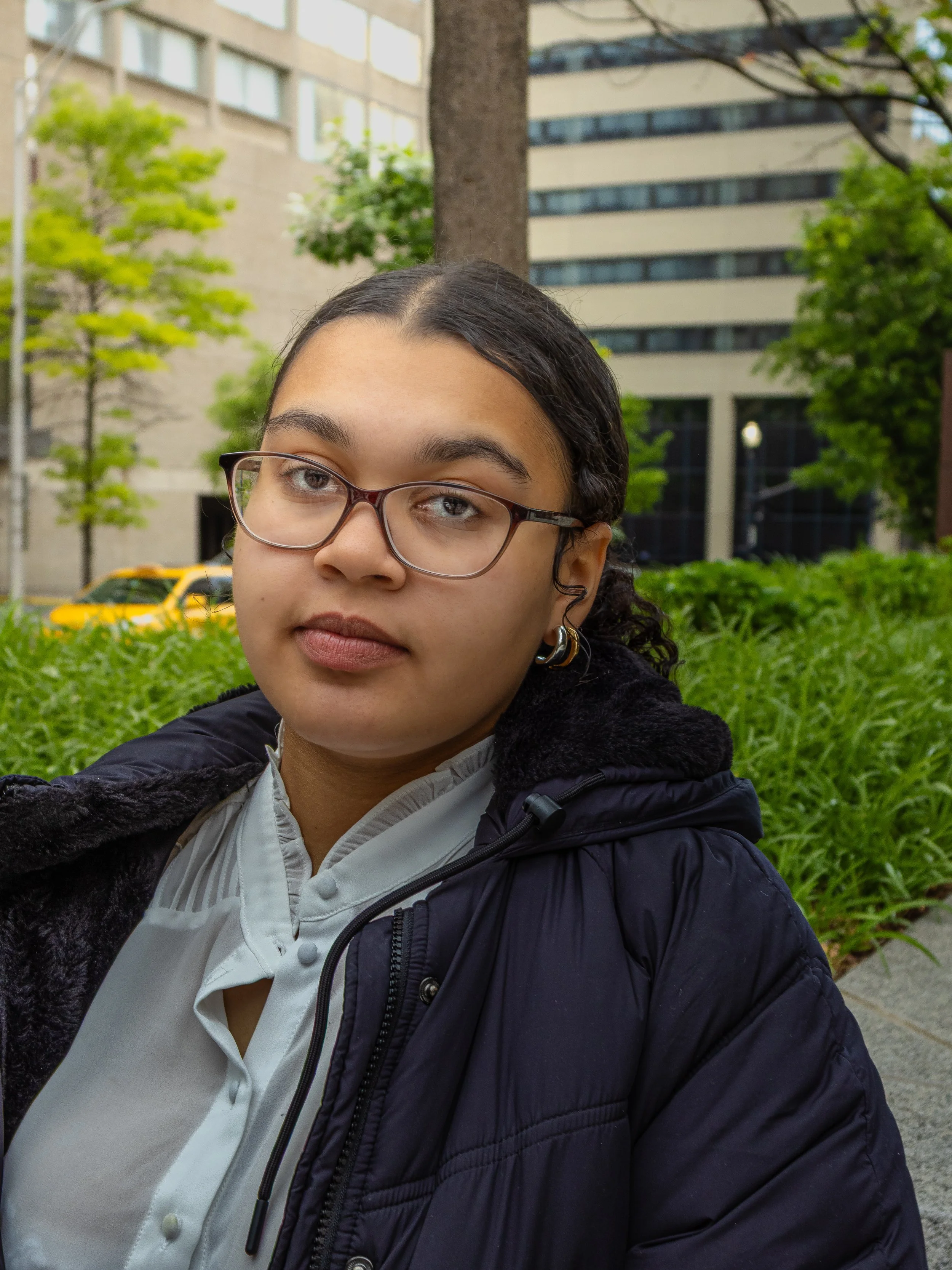 A young woman with glasses and dark hair pulled to the side, standing outdoors in a city environment with trees, buildings, and a yellow taxi visible in the background.